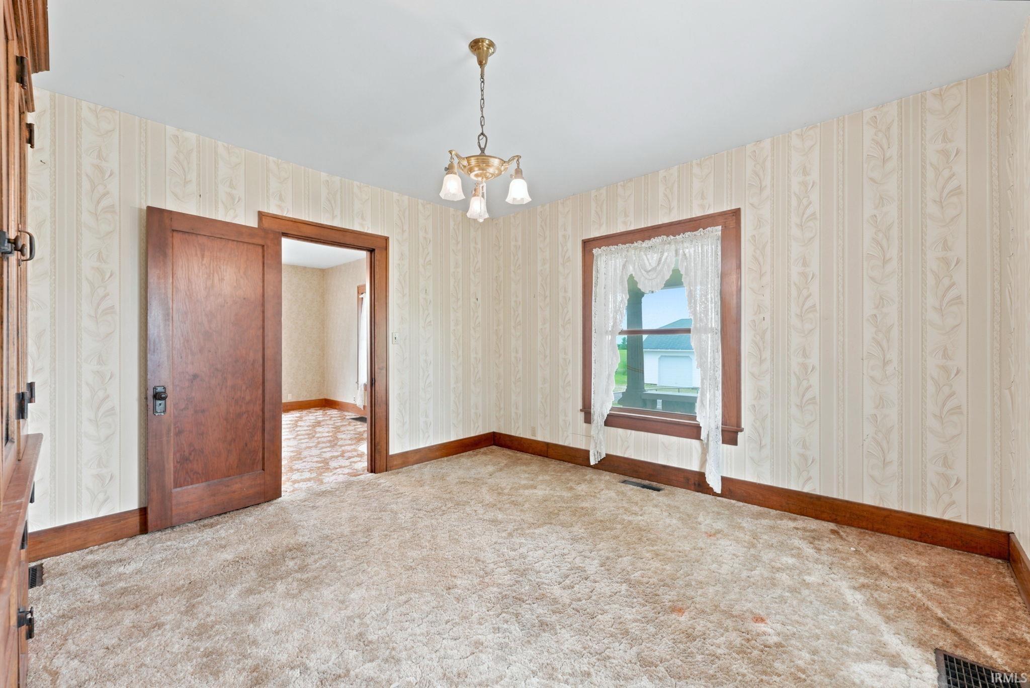 Room featuring patterned wallpaper, carpeting, a ceiling-mounted chandelier, and wood trim around the window and door frame
