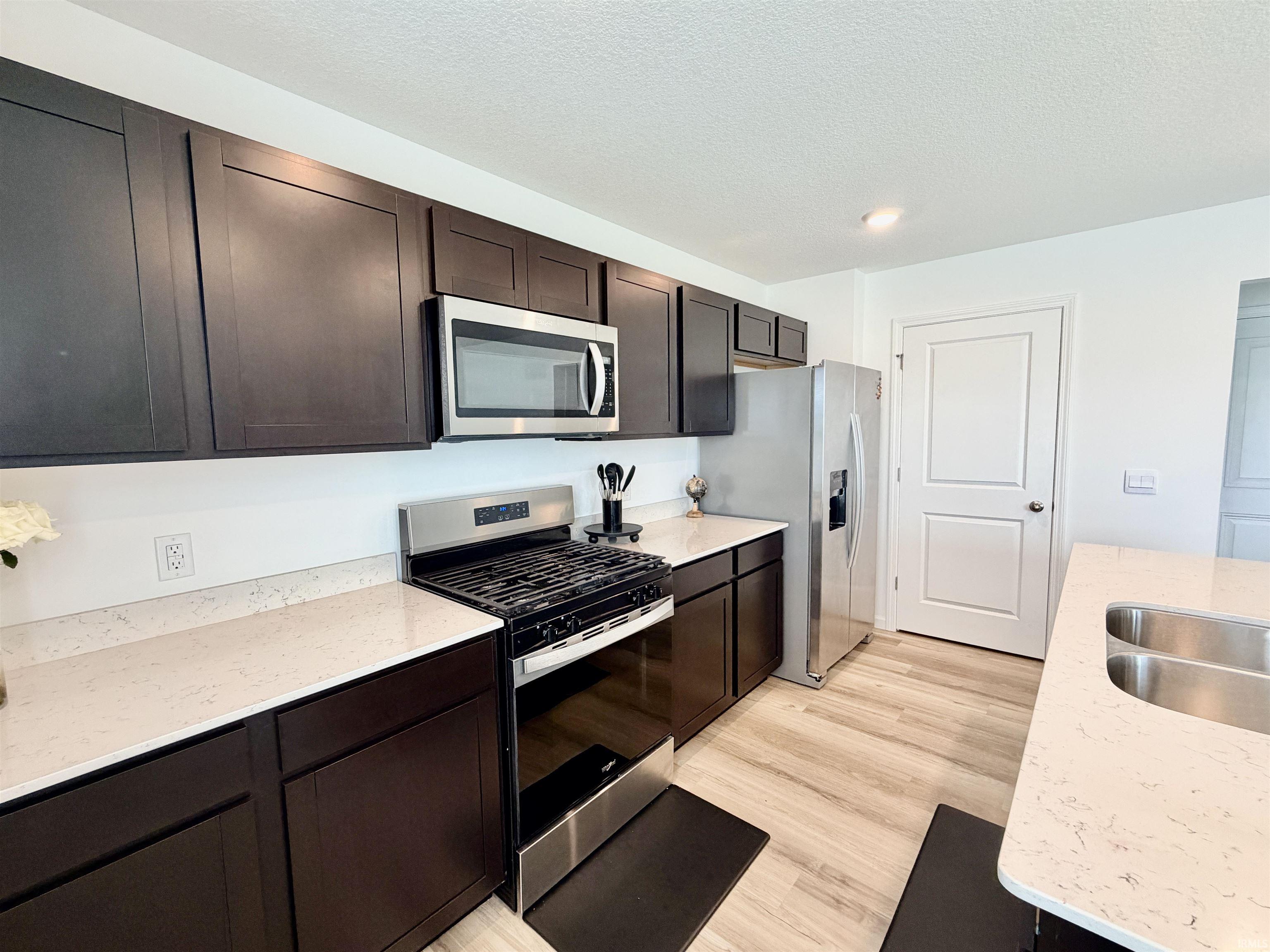 Kitchen featuring dark wood-finish cabinetry, light-toned countertops, stainless steel appliances, a double basin sink, and light wood-finish flooring
