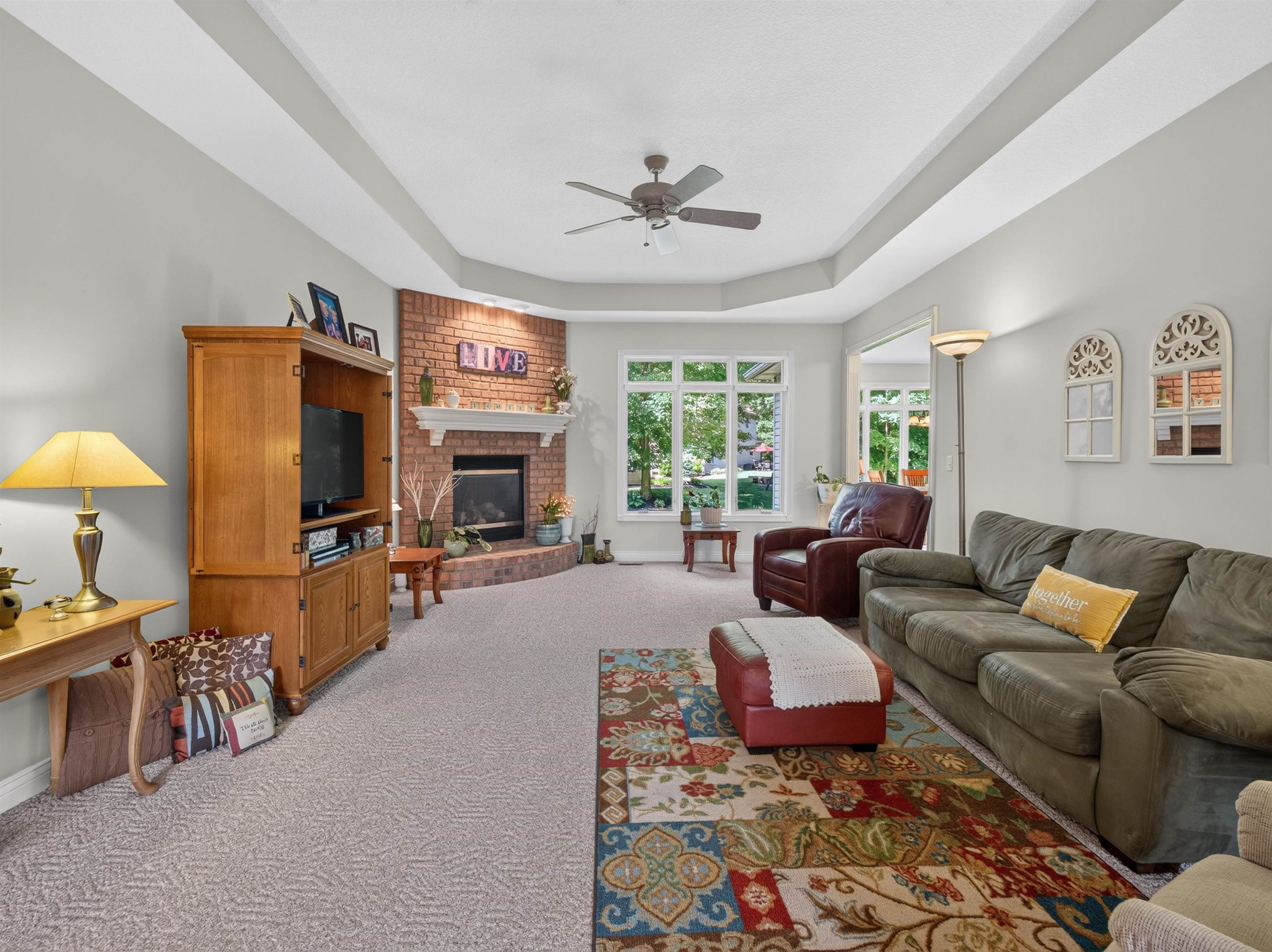 Spacious living area featuring a tray ceiling with a ceiling fan, a brick fireplace with a white mantel, and large windows
