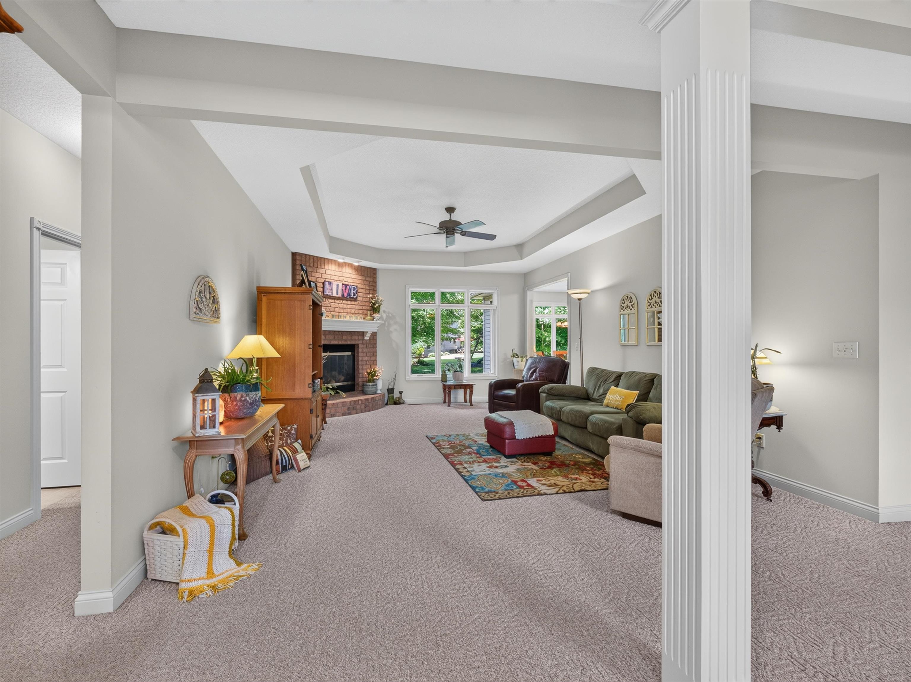 Spacious living area featuring a tray ceiling with a ceiling fan, a corner brick fireplace with a white mantel, and large windows
