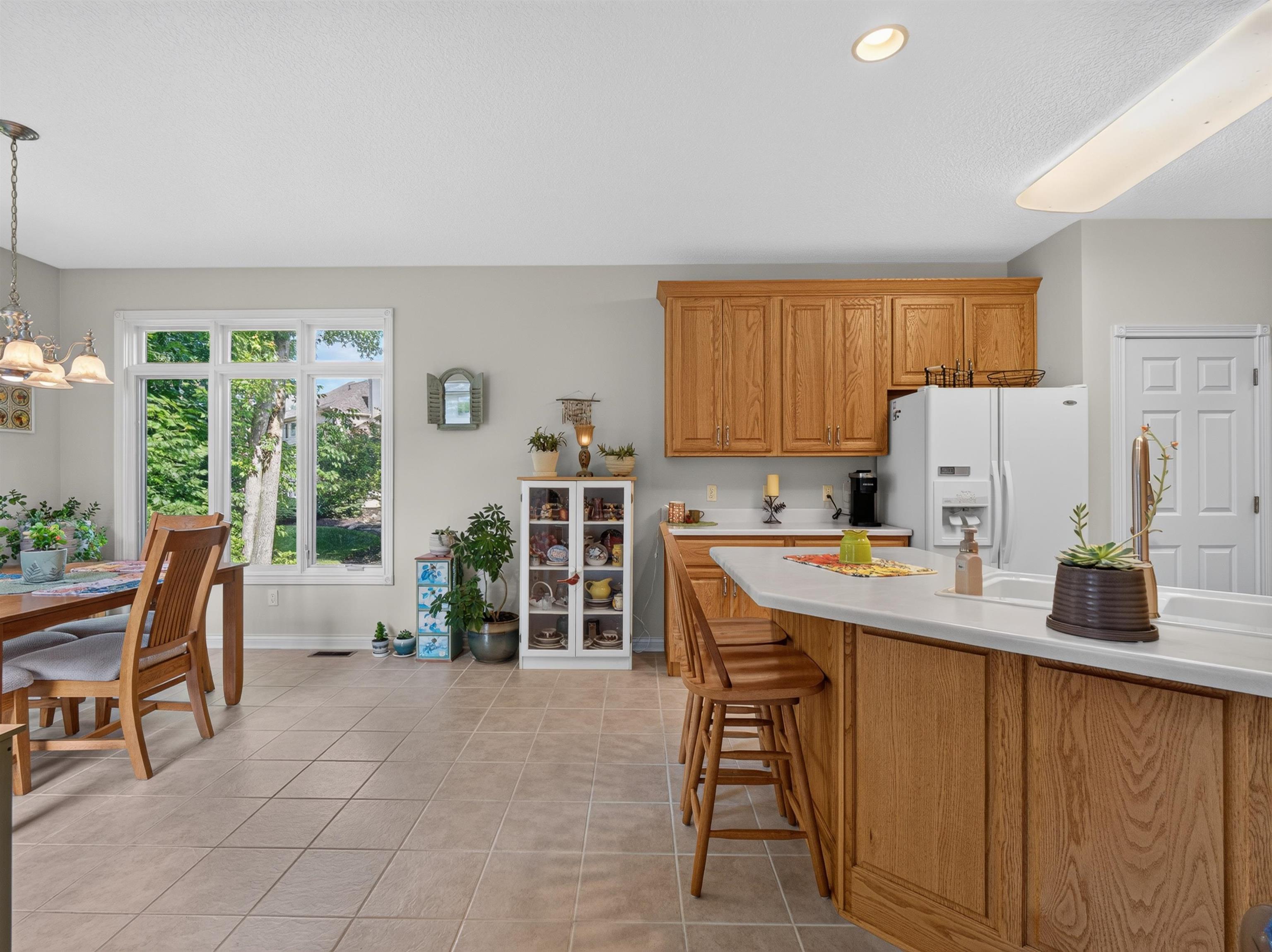Kitchen and dining area featuring light-toned tile flooring, honey-toned wood cabinetry, a white countertop island with seating, a large window, and recessed lighting