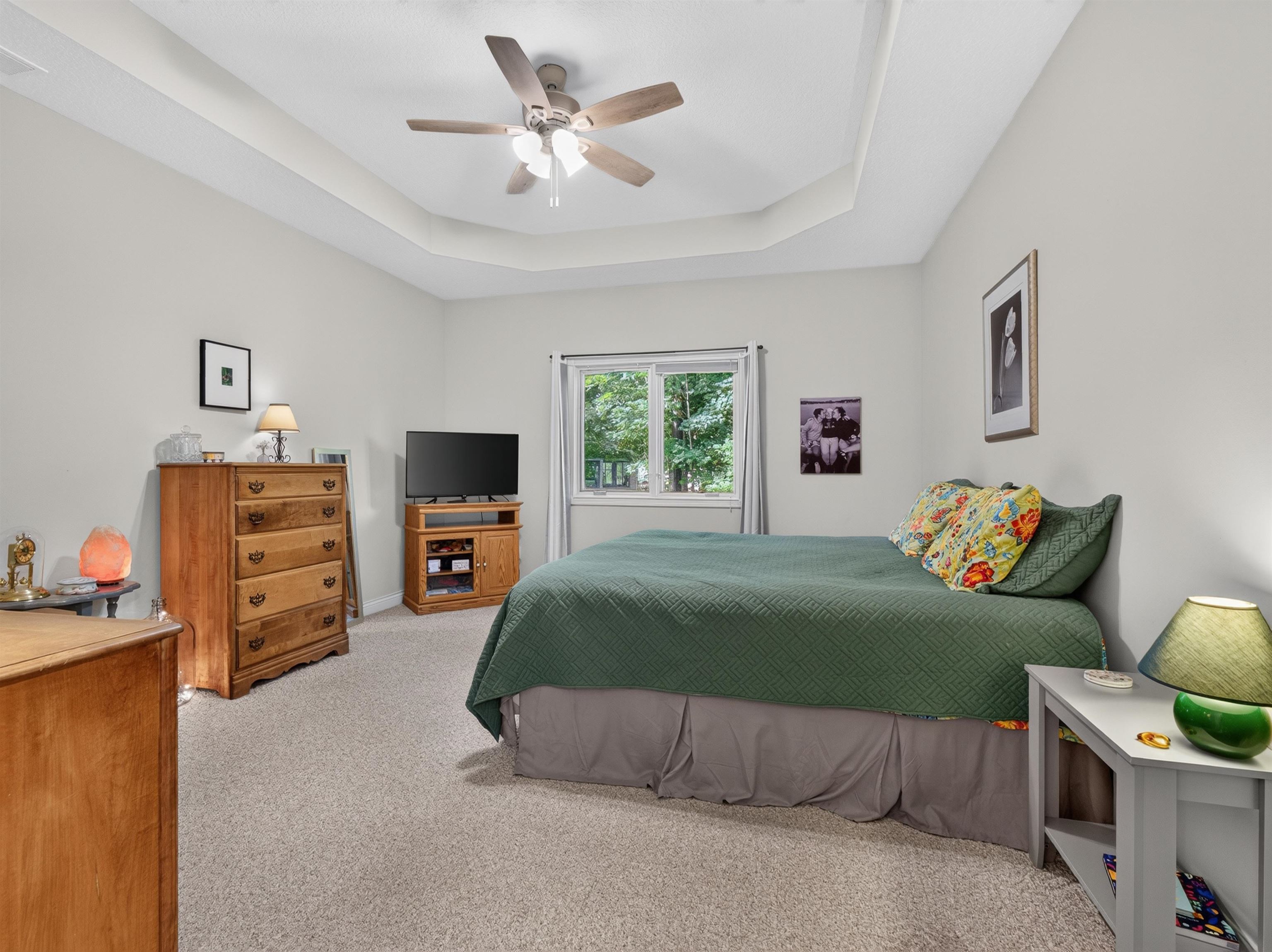 Spacious room featuring a tray ceiling with a ceiling fan, light-toned carpeting, and a window with a wooded backdrop