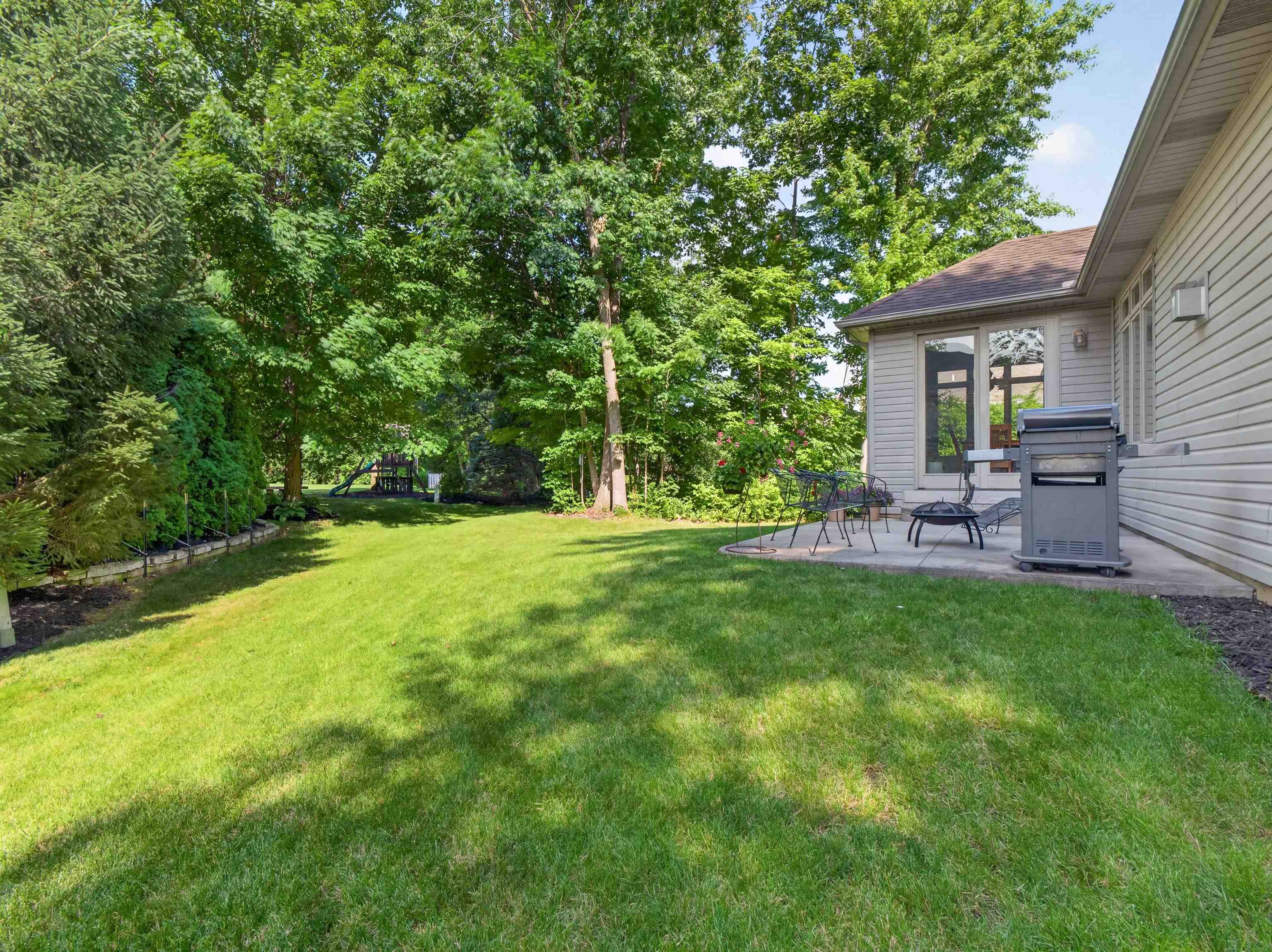 Expansive lawn area featuring a curved concrete patio, mature perimeter trees, vinyl siding, and an attached outdoor lighting fixture