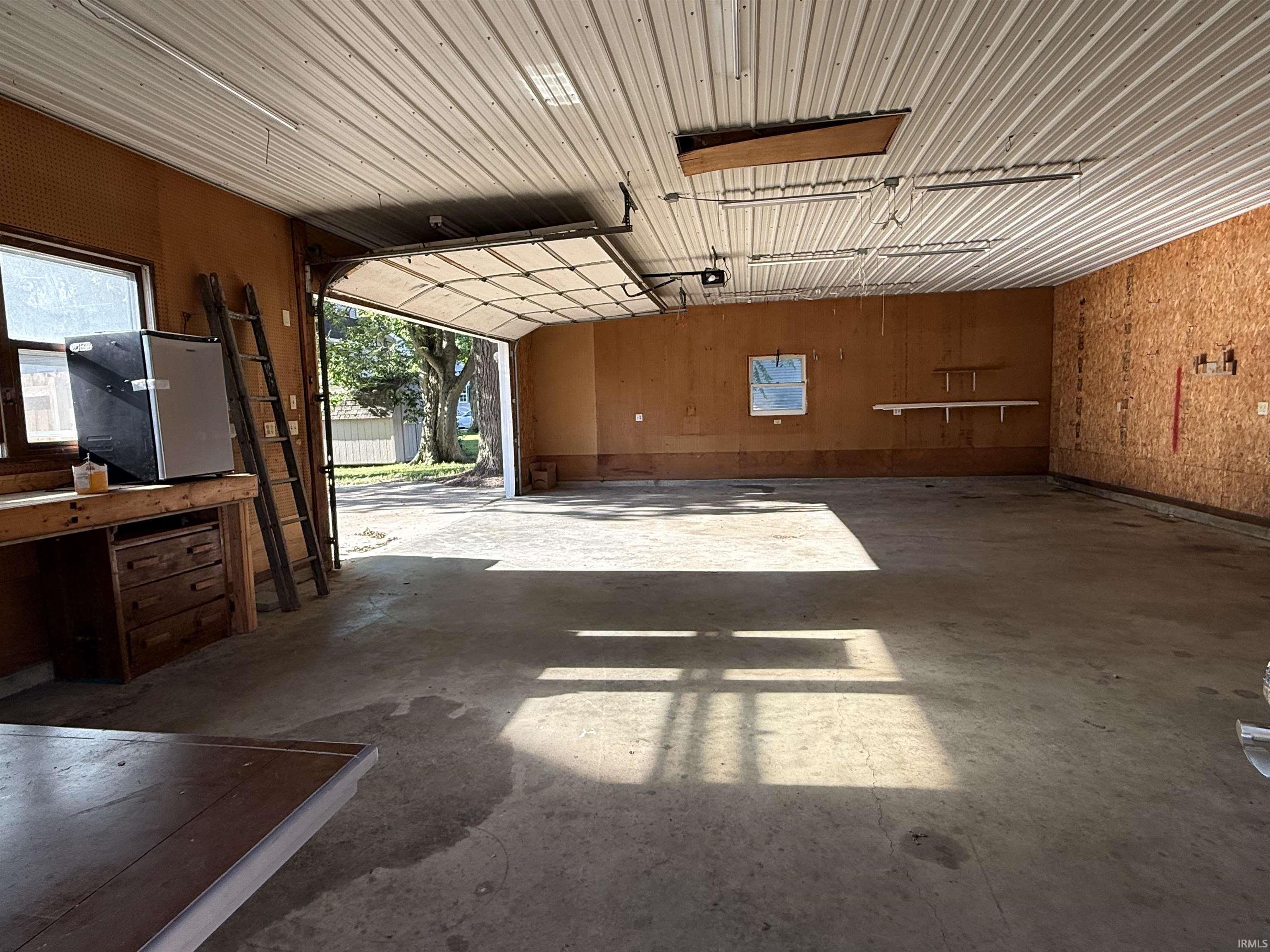 Spacious garage featuring a concrete floor, roll-up garage door, metal corrugated ceiling, and built-in workbench with drawers