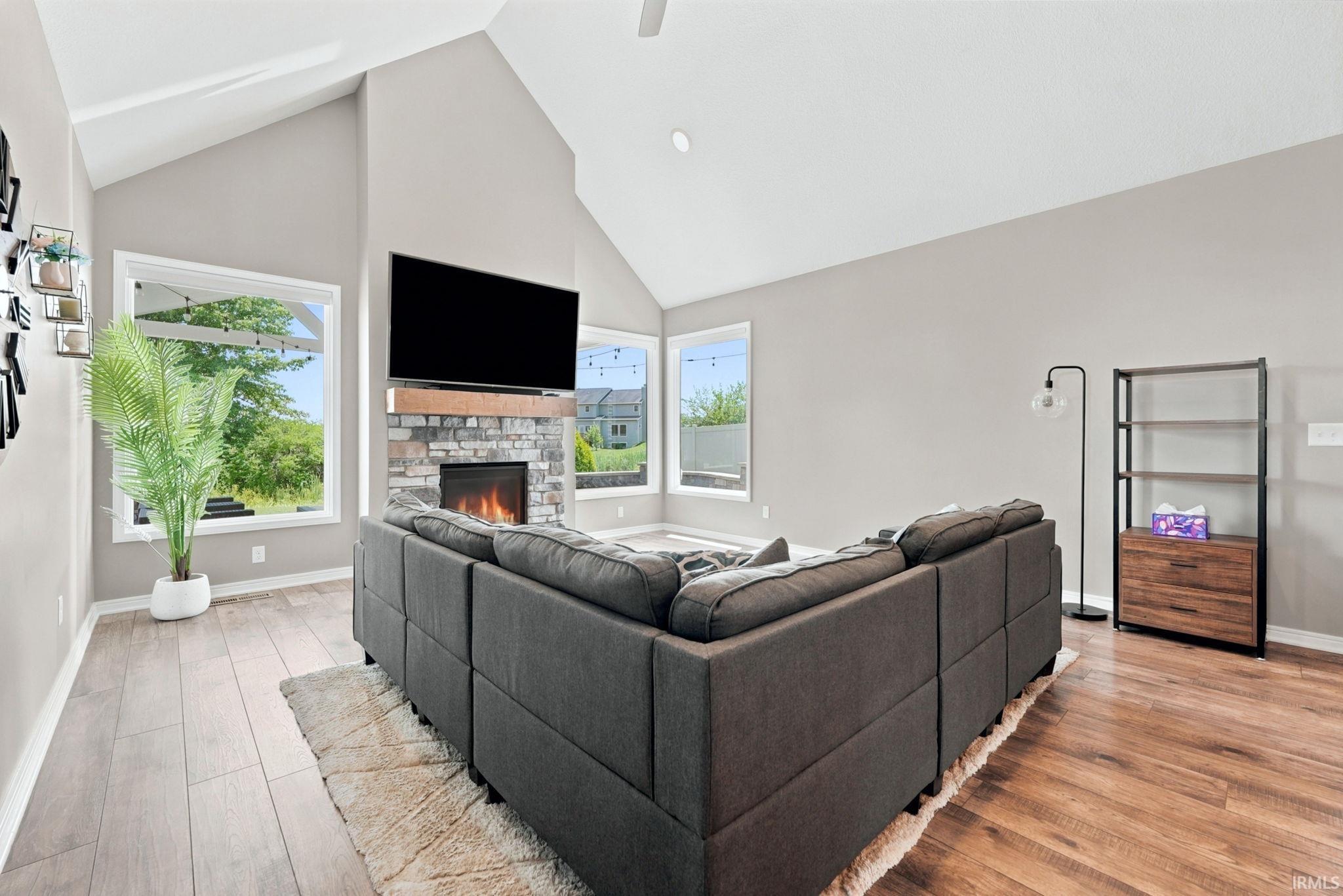 Vaulted ceilings accentuate this living space, featuring a stone fireplace with a wood mantle, large windows, and contrasting wood-finish flooring