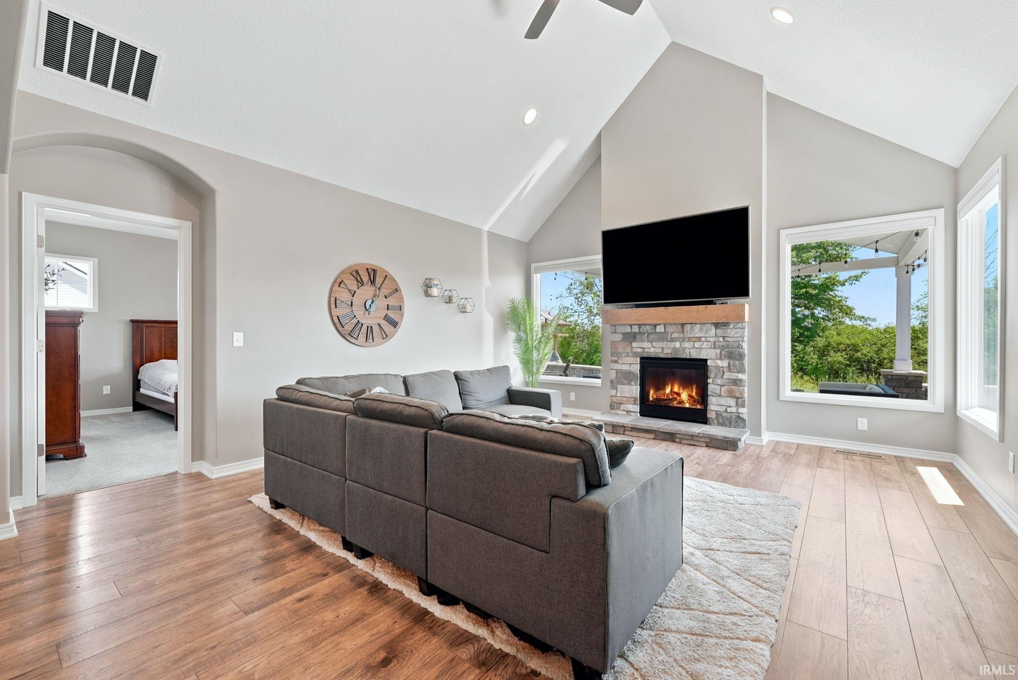 Spacious living area featuring vaulted ceilings, wood-finish flooring, a stone fireplace with a wood mantle, and large windows