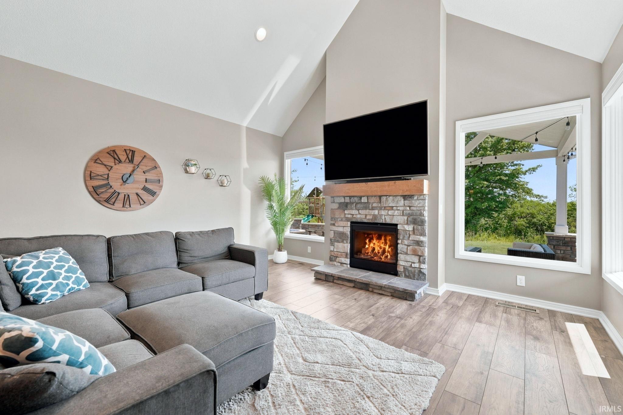 Vaulted ceiling living area featuring a stone-faced fireplace with a wood mantel, wood-finish flooring, and large windows
