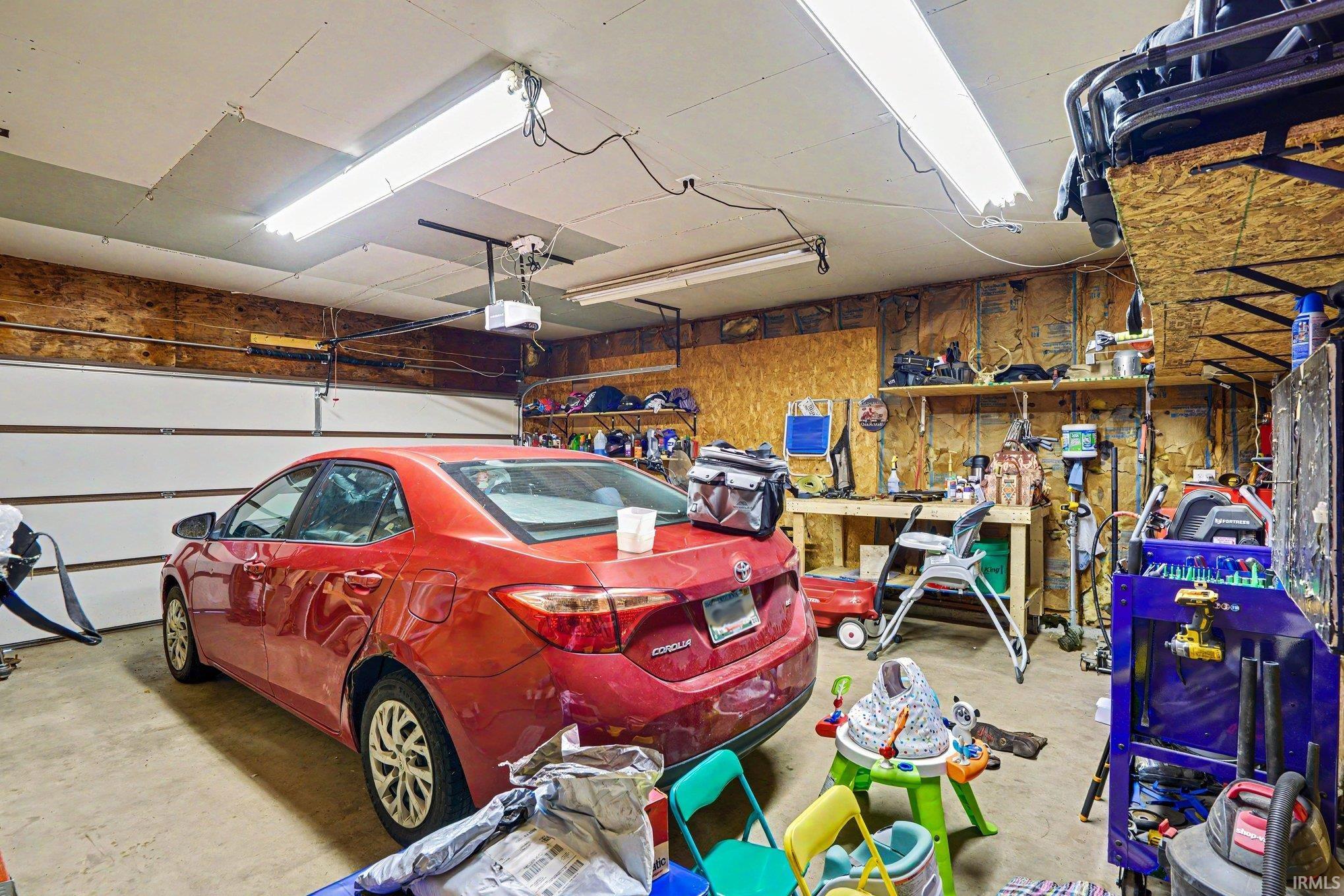 Attached garage featuring two overhead doors, exposed plywood walls, built-in shelving, and overhead fluorescent lighting