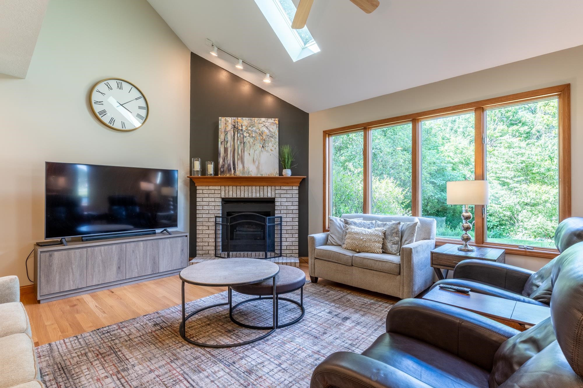 Vaulted ceiling living space featuring a brick fireplace with a wood mantle, hardwood flooring, a skylight, and large picture windows