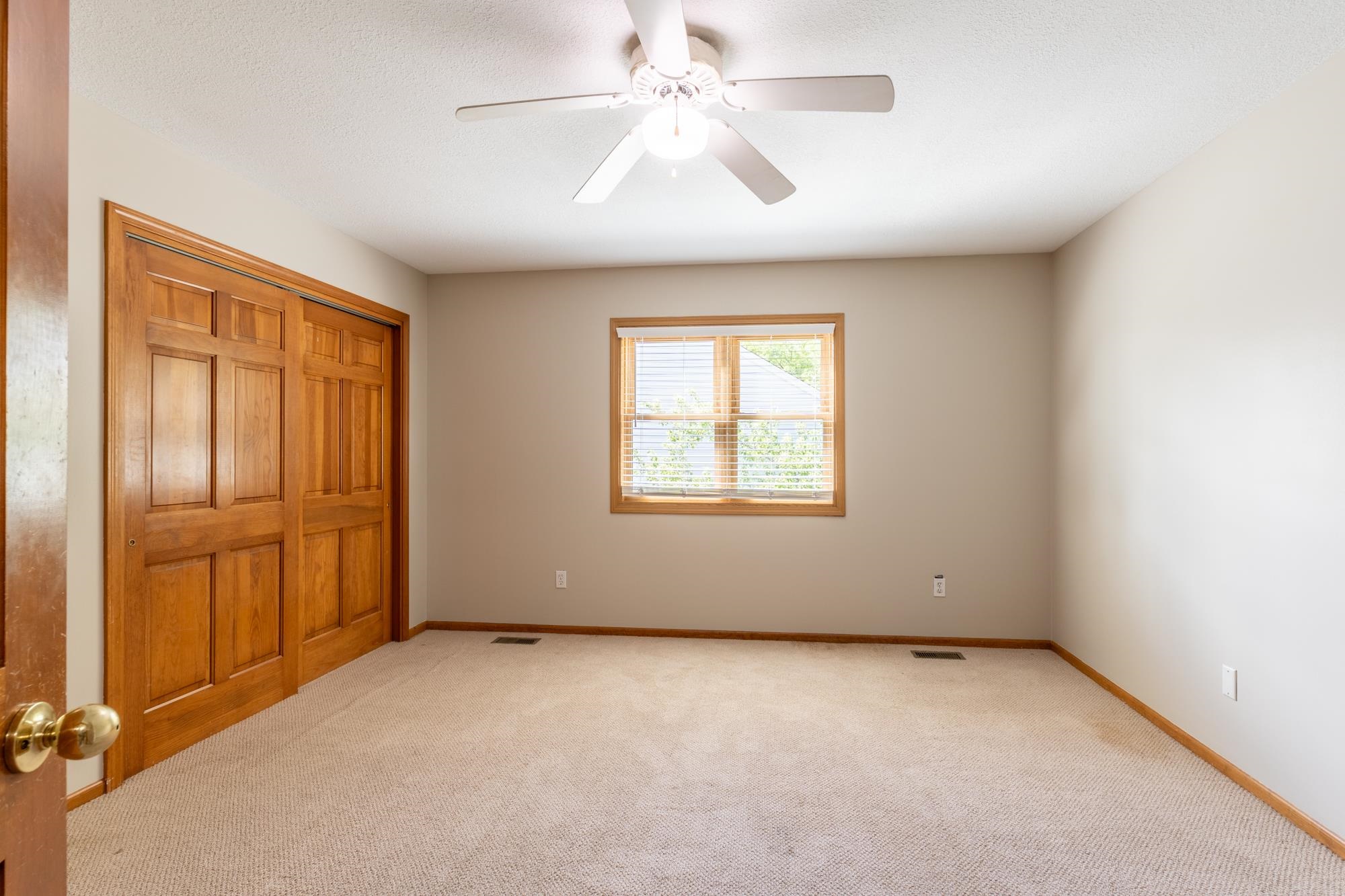 Neutral-toned room featuring a ceiling fan, light-colored carpeting, a window with wood-finish trim, and sliding wood-finish closet doors