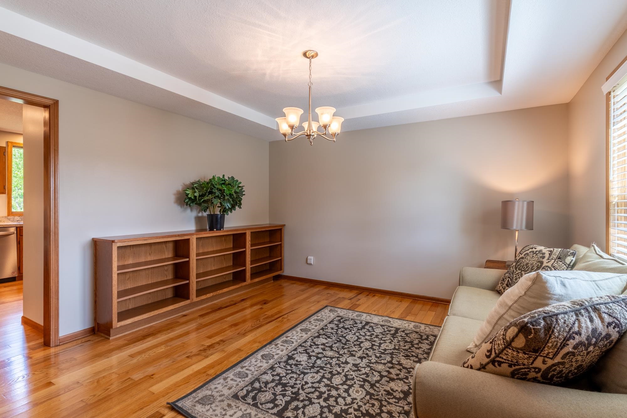 Tray ceiling with chandelier lighting, wood-finish flooring, built-in shelving, wood door trim, and window with blinds