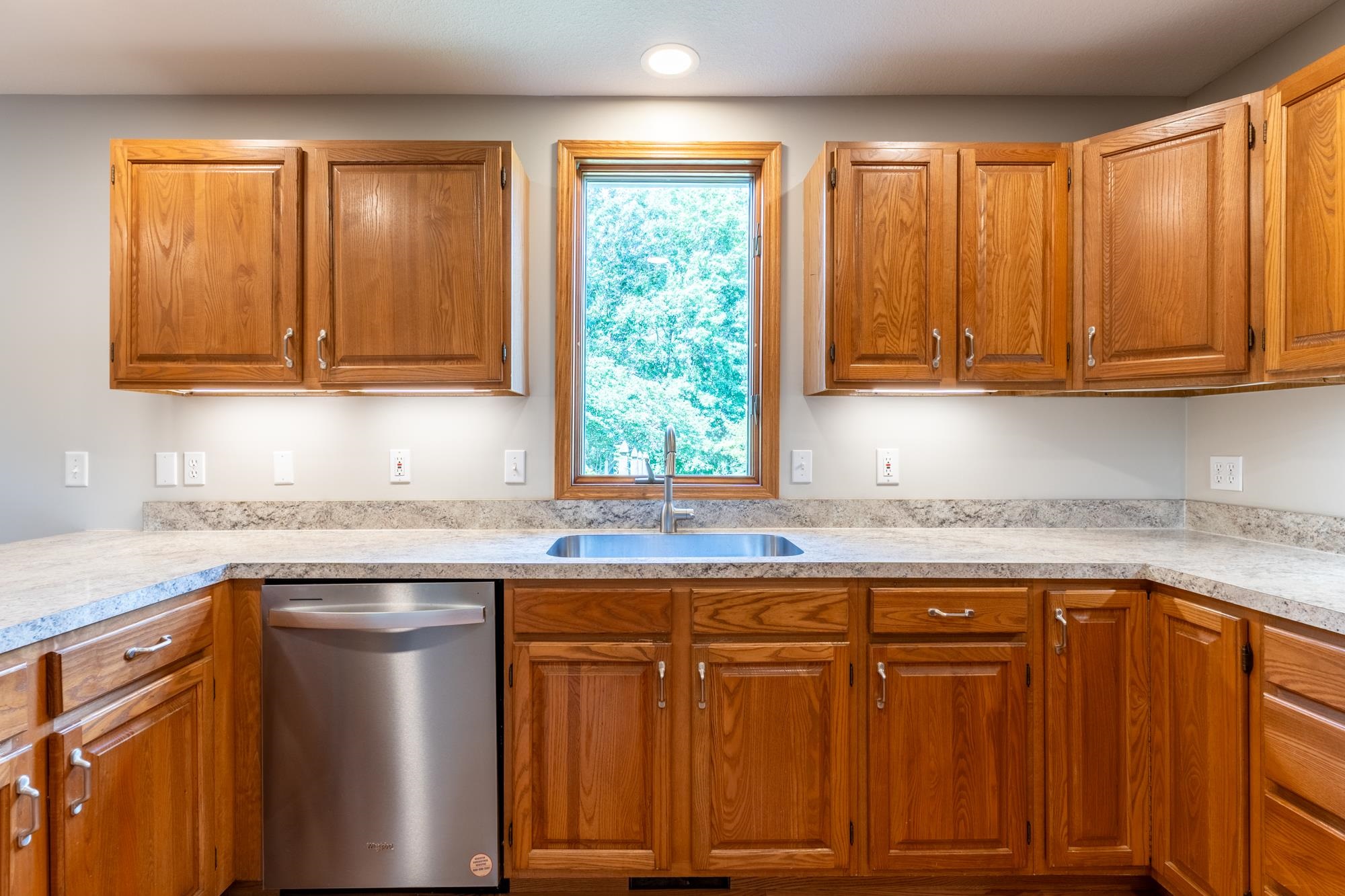 Kitchen featuring wood cabinetry, a stainless steel dishwasher, an undermount sink, a window with exterior visibility, and speckled countertops