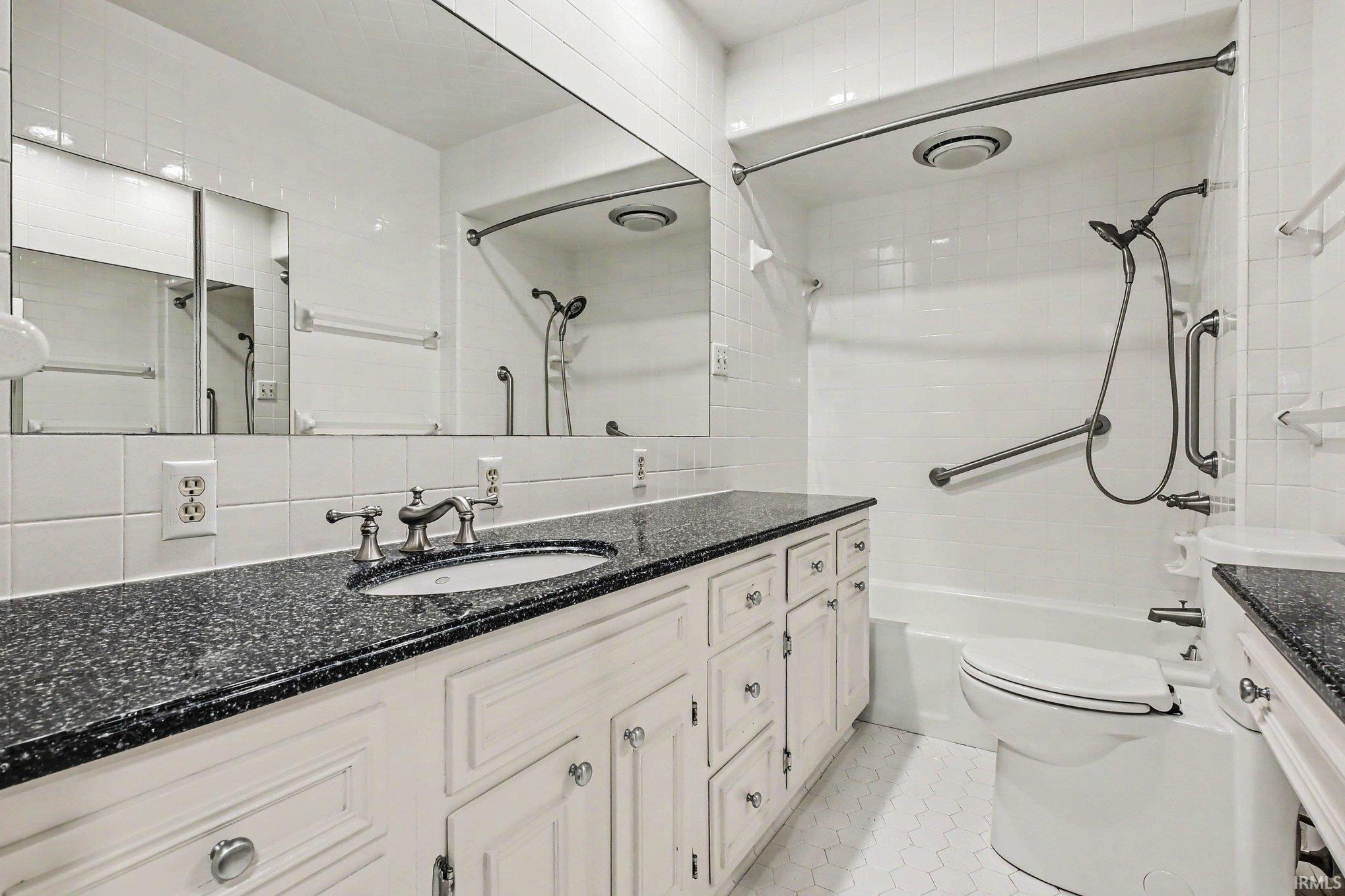 Bathroom featuring a vanity with black countertops, white cabinetry with silver hardware, a large wall-mounted mirror, and a white tiled shower-tub combination with grab bars