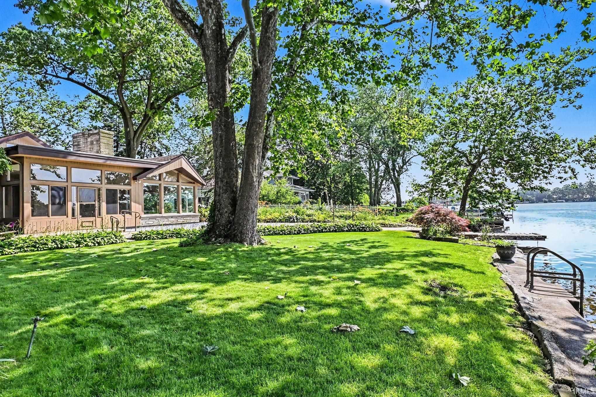 Waterfront property featuring expansive window walls, mature trees, a manicured lawn, gardens and a private dock and pier with water access