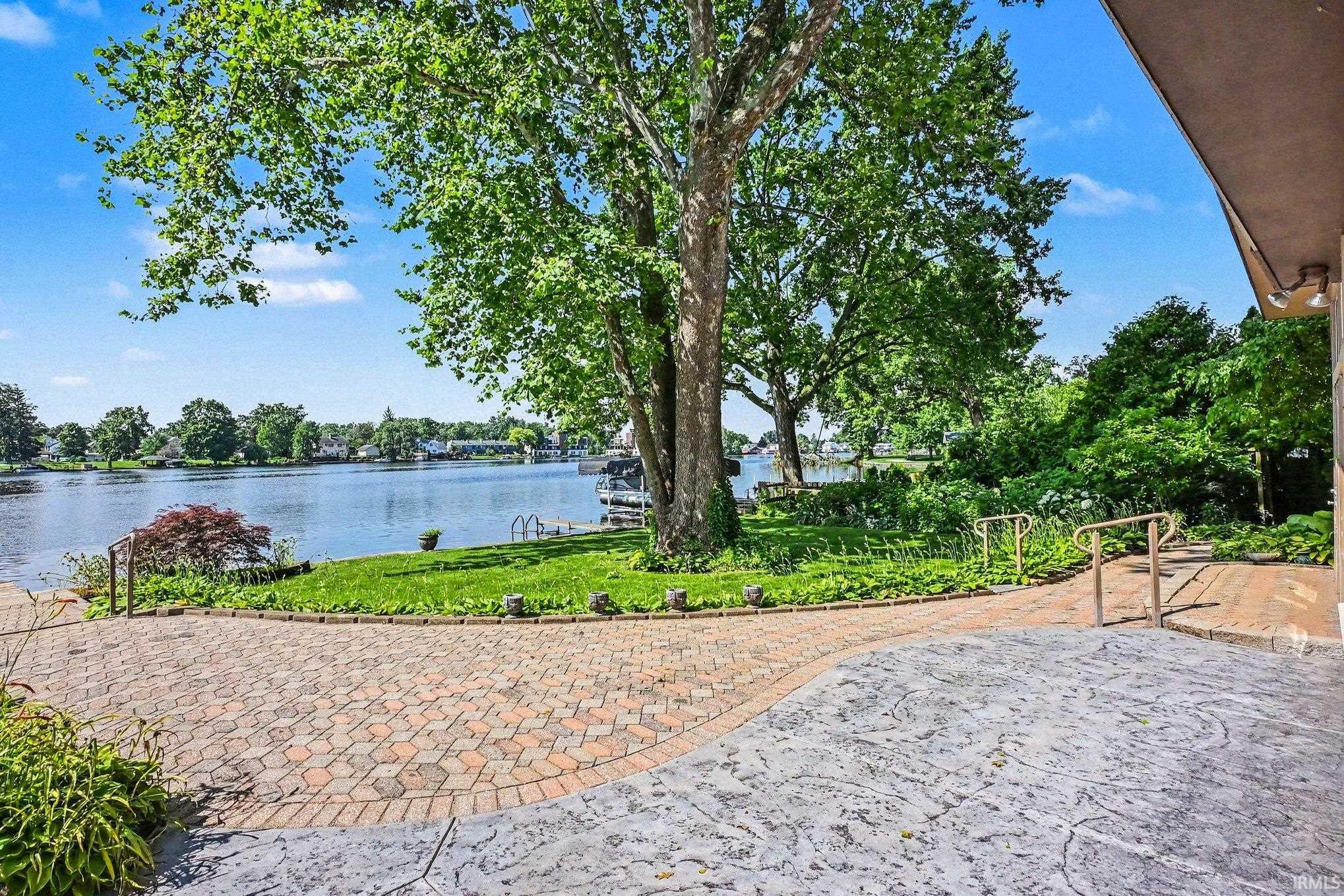 Waterfront property featuring a flagstone and brick patio and walkway, mature trees, manicured lawn, gardens, and steps with railings leading to the water