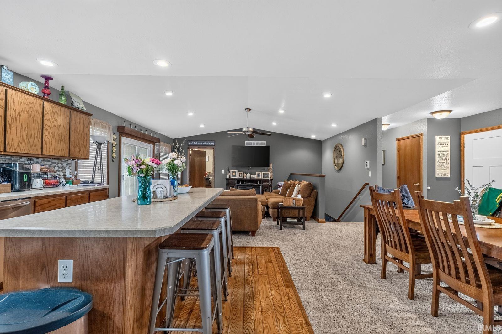 Open-concept living area featuring a kitchen island with seating, recessed lighting, vaulted ceilings, a carpeted living space, and wood-finish flooring