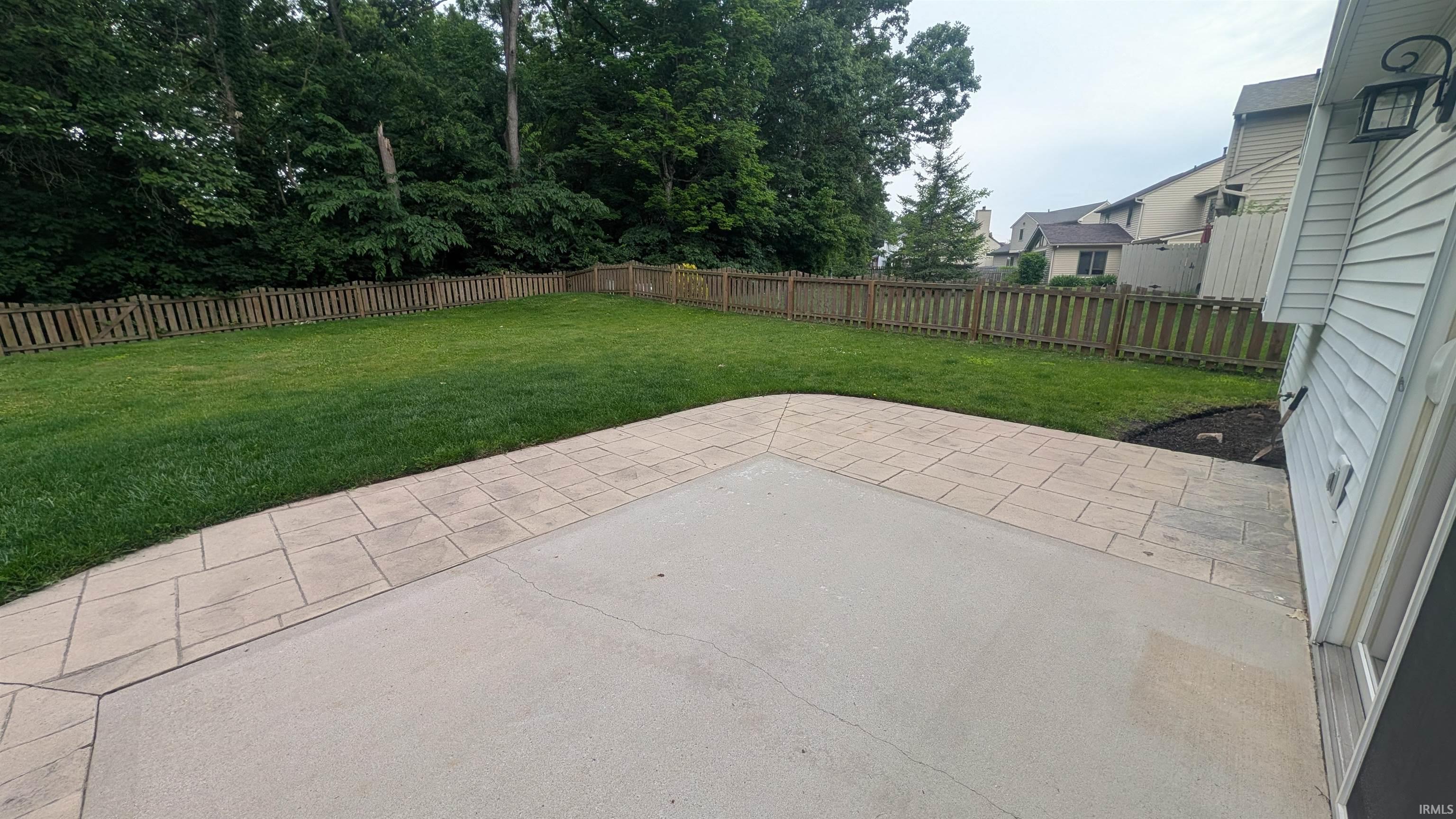 Expansive backyard featuring a large concrete patio with decorative paver edging, a manicured lawn, and a perimeter wooden fence