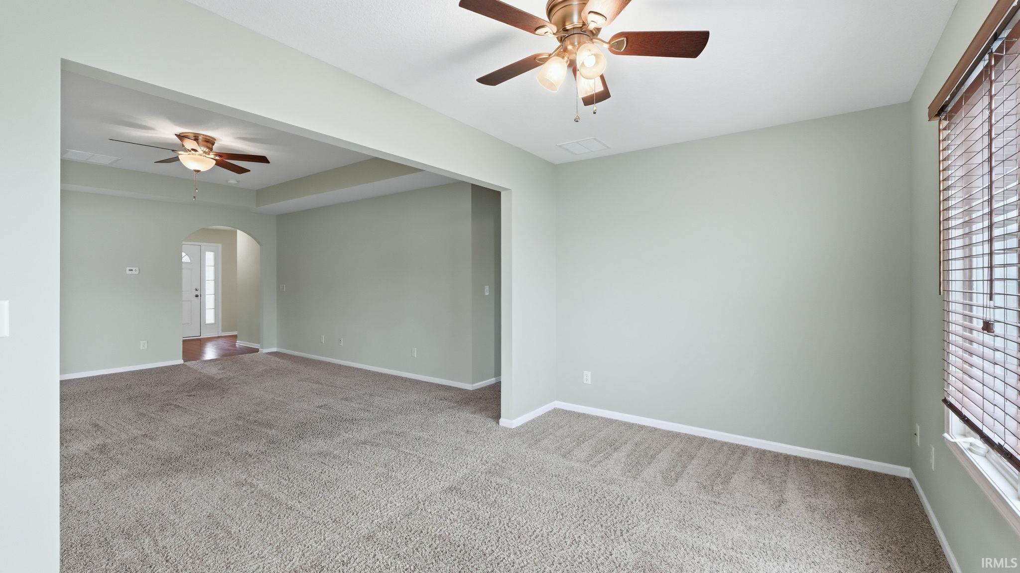 Expansive carpeted room featuring a ceiling fan with integrated lighting, window with horizontal blinds, and a wide cased opening to an additional area