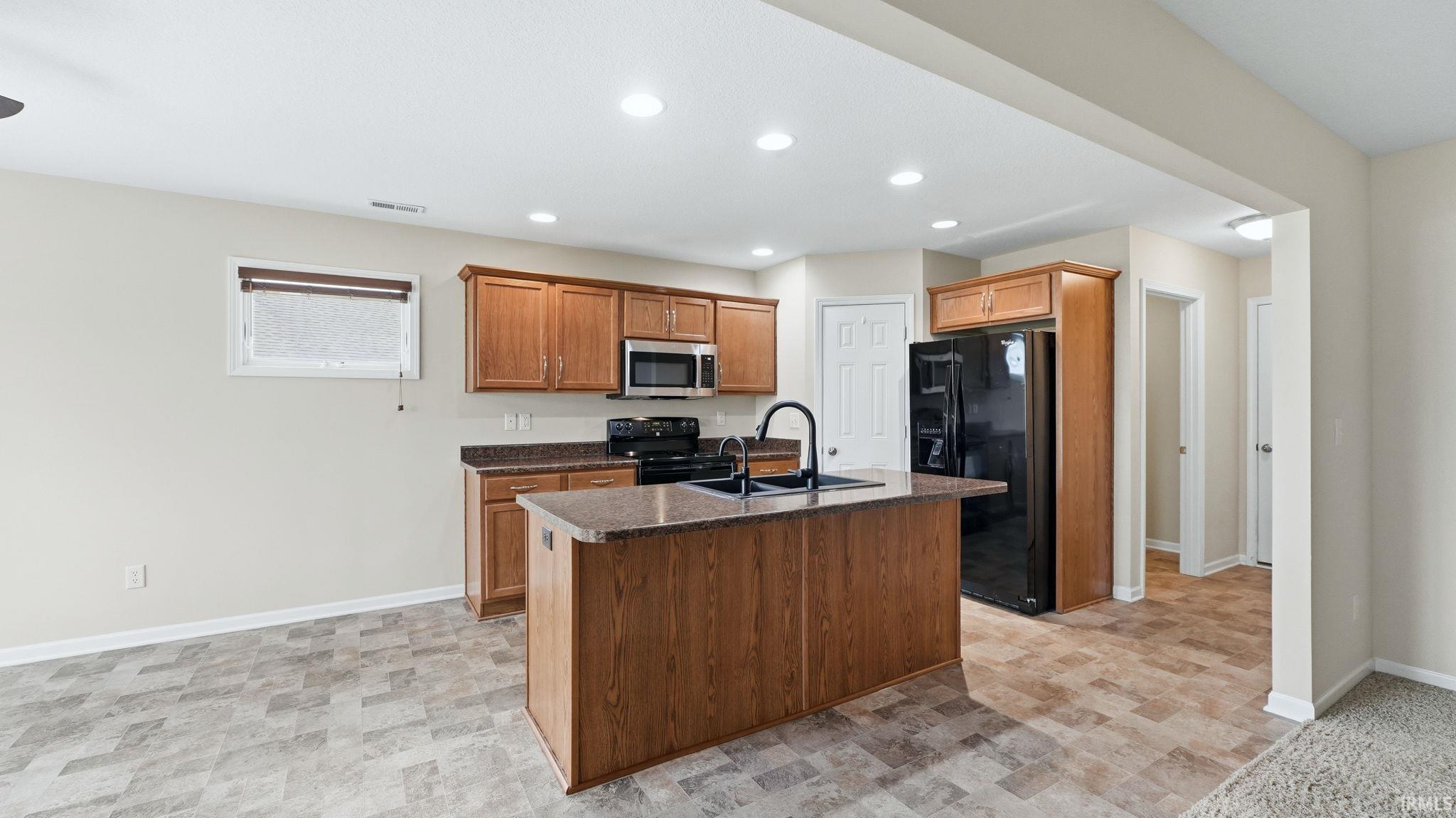 Open-concept kitchen featuring wood-finish cabinetry, black appliances, a central island with a sink and faucet, and a stone-finish countertop