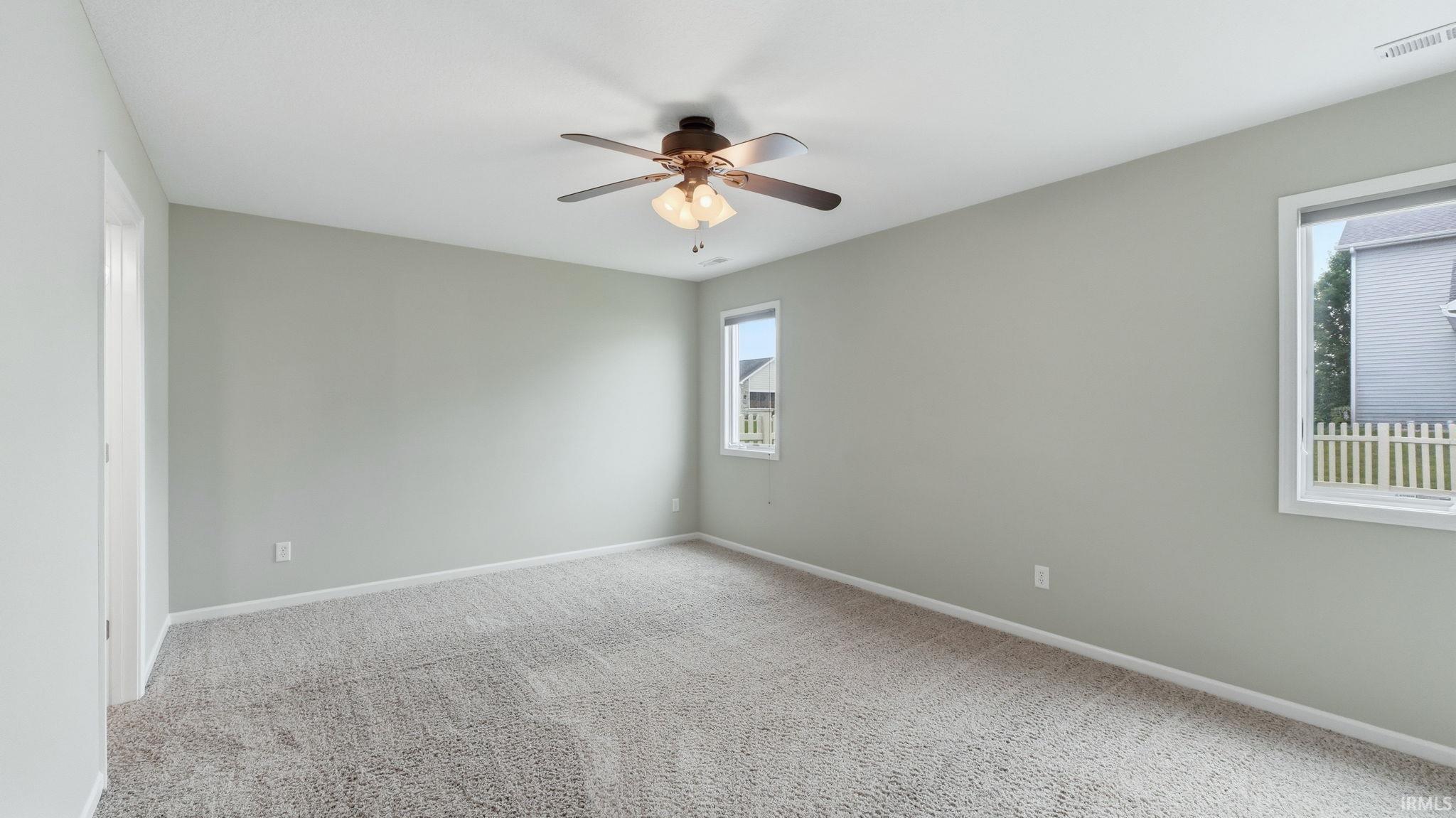 Spacious interior room featuring light-toned walls, textured gray carpeting, and a ceiling fan with integrated lighting
