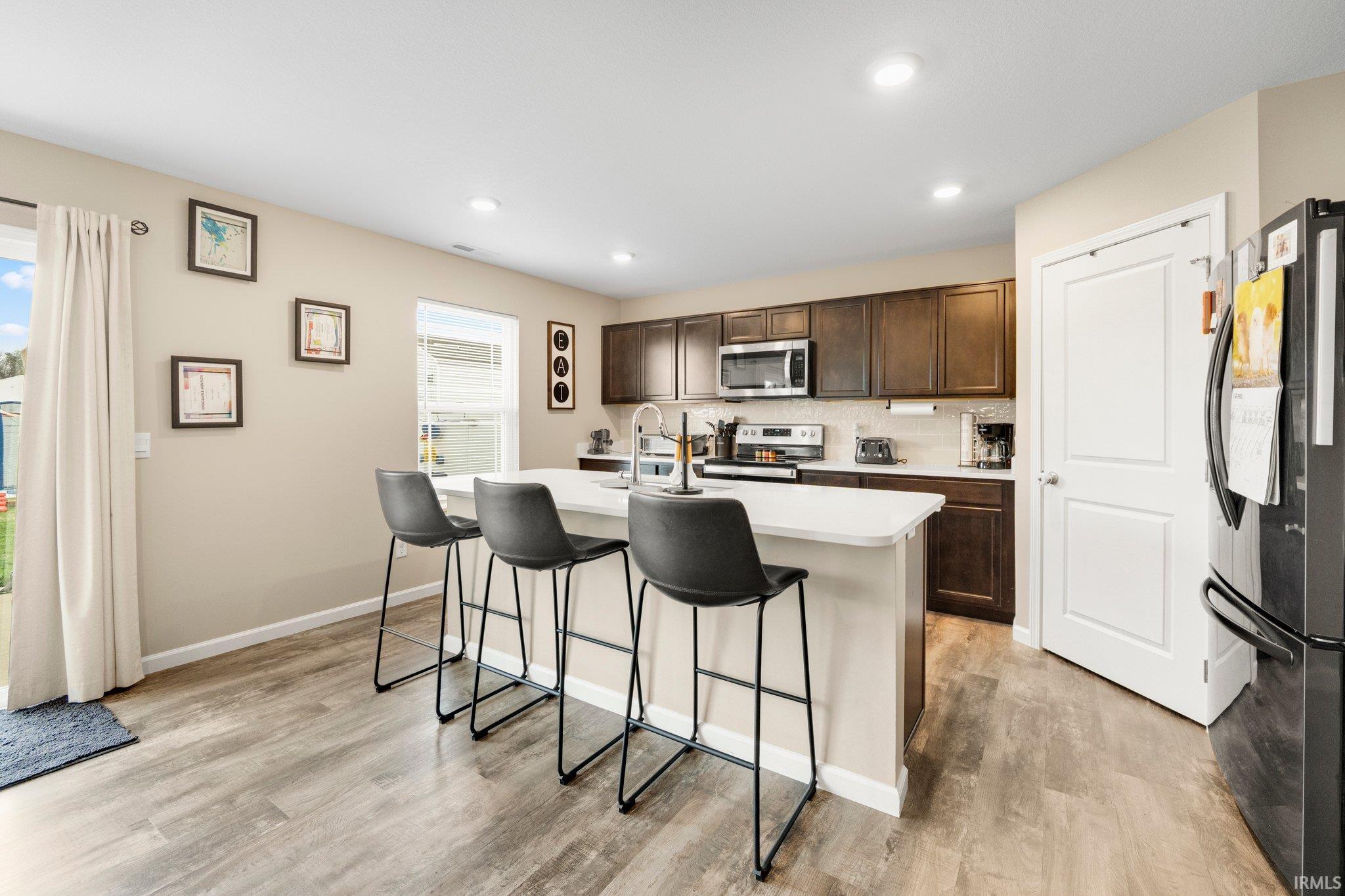 Modern kitchen featuring an island with a light-toned countertop, wood-finish flooring, dark wood cabinetry, stainless steel appliances, and recessed lighting