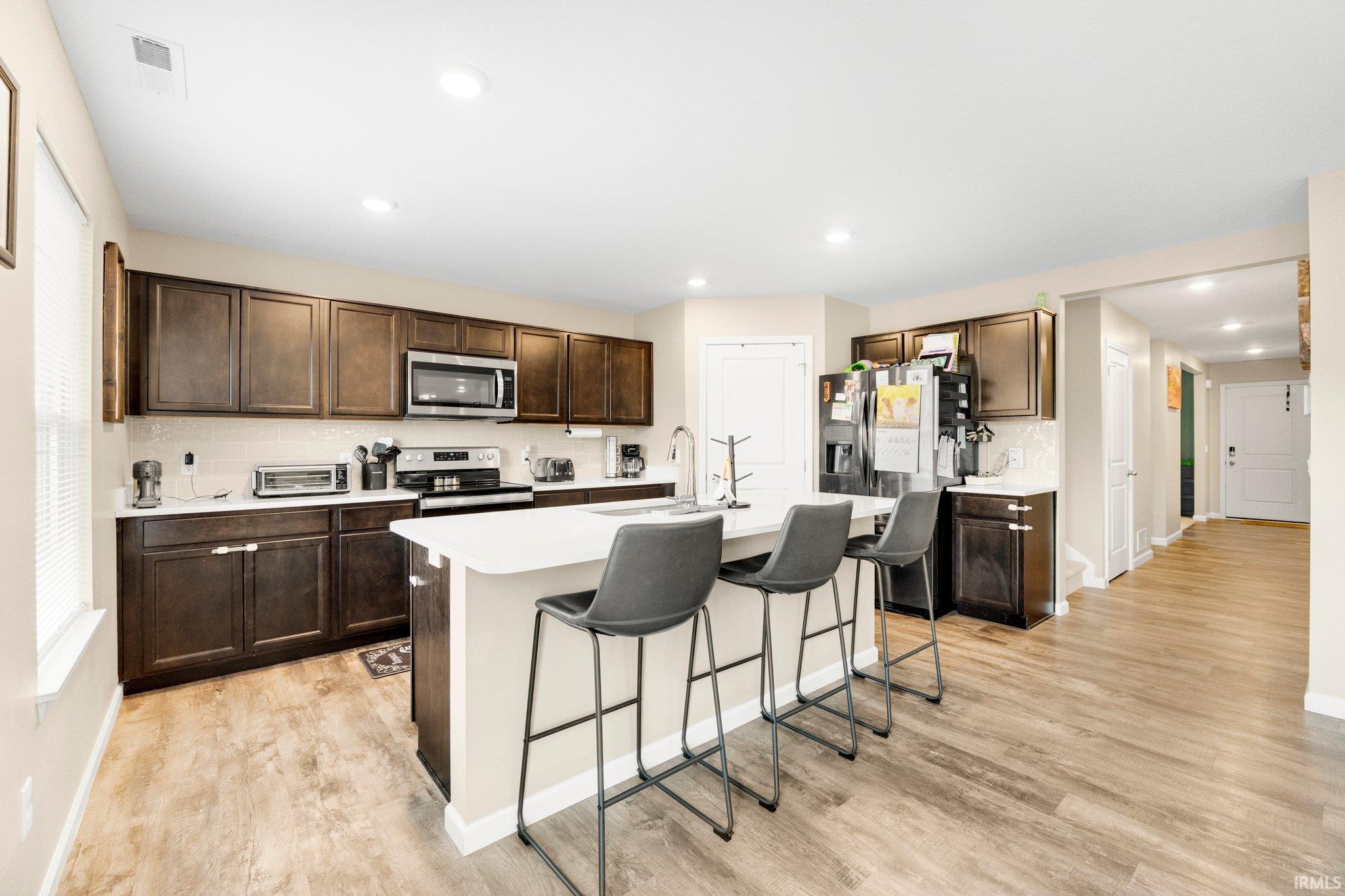 Open-concept kitchen featuring an island with seating, wood-finish flooring, dark wood cabinetry, white countertops, and stainless steel appliances