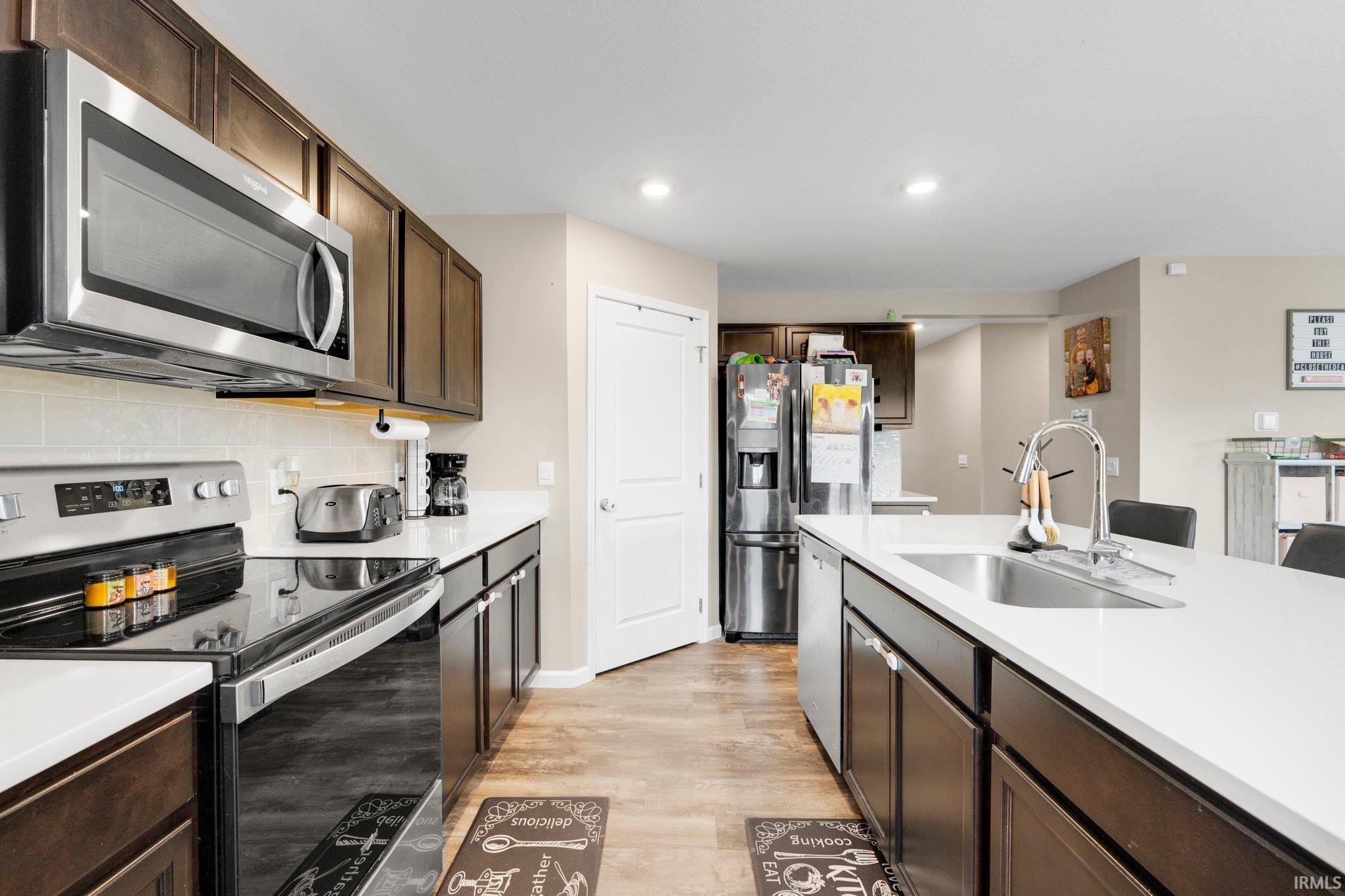 Kitchen featuring an expansive center island with an undermount sink, stainless steel appliances, dark wood-finish cabinetry, light countertops, and wood-finish flooring