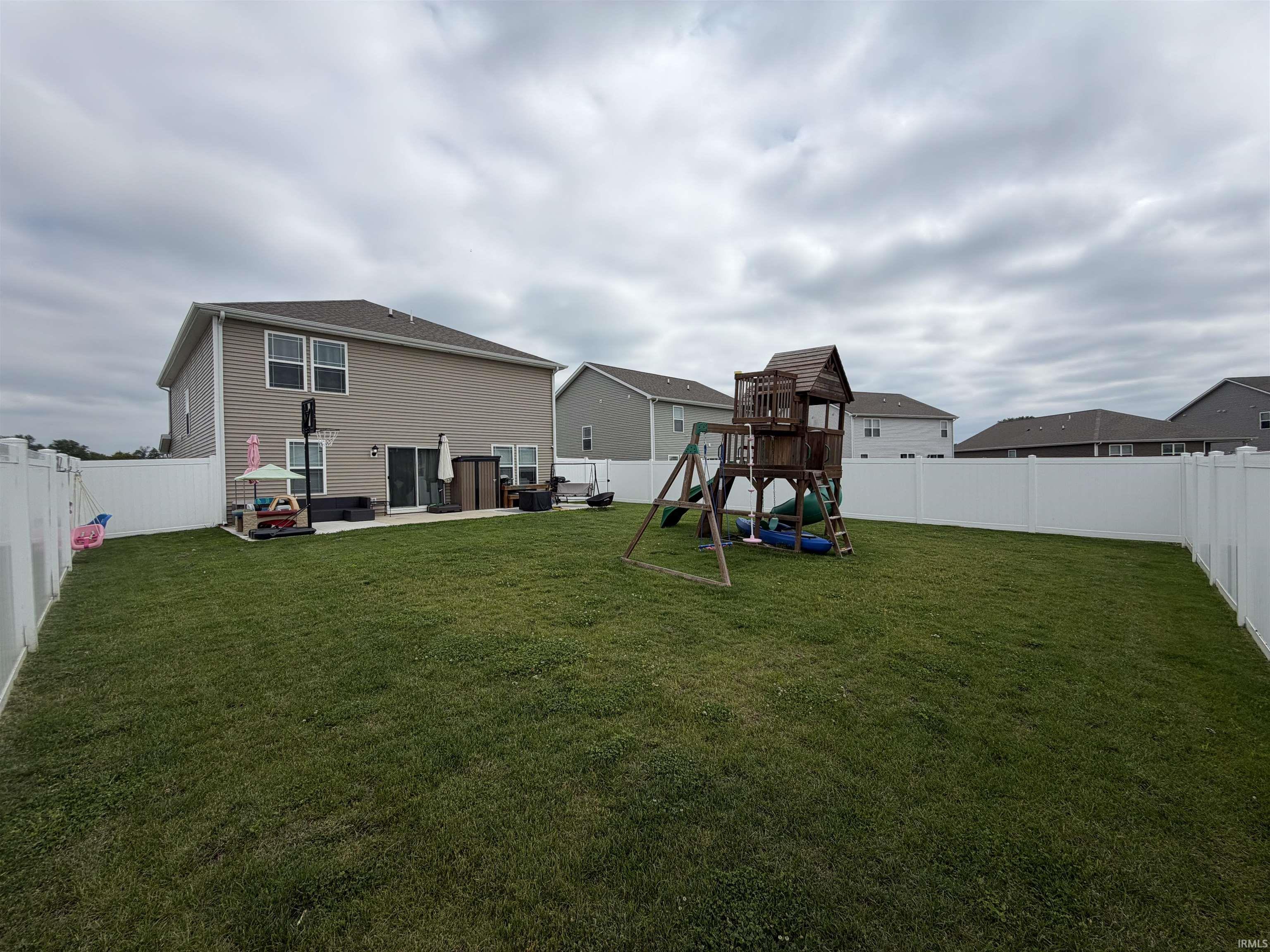 Expansive backyard featuring a manicured lawn, full white vinyl fencing, a concrete patio, and a two-story residence with tan siding and white trim