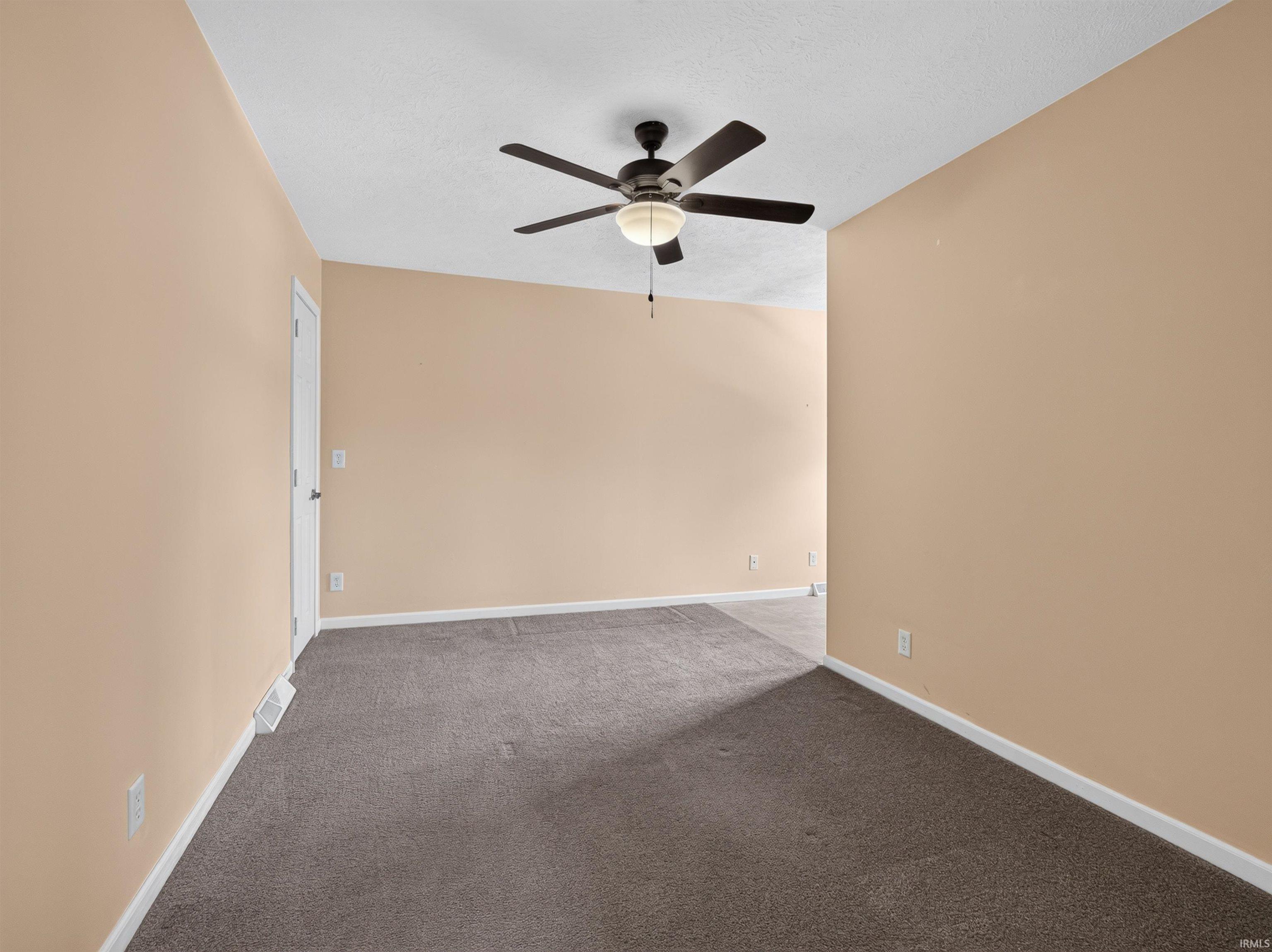 Spacious interior room featuring a ceiling fan with integrated lighting, textured ceilings, a warm neutral wall color, and dark gray carpeting