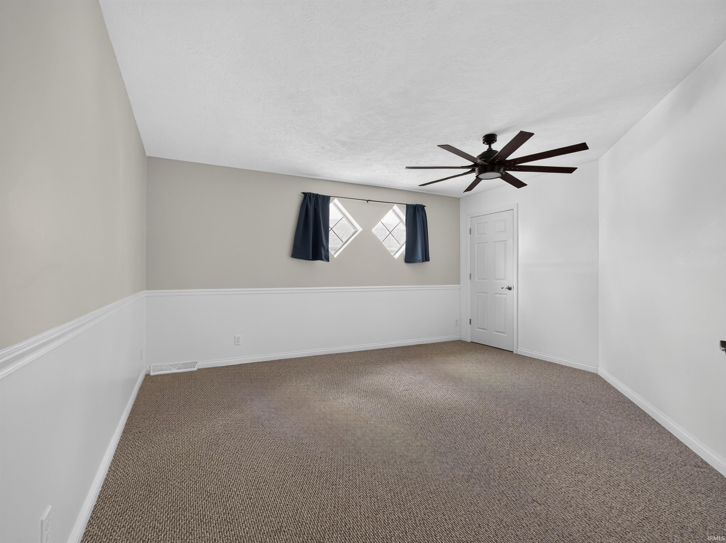 Spacious carpeted room featuring a modern ceiling fan, two diamond-pane windows, wainscoting, and a white paneled door