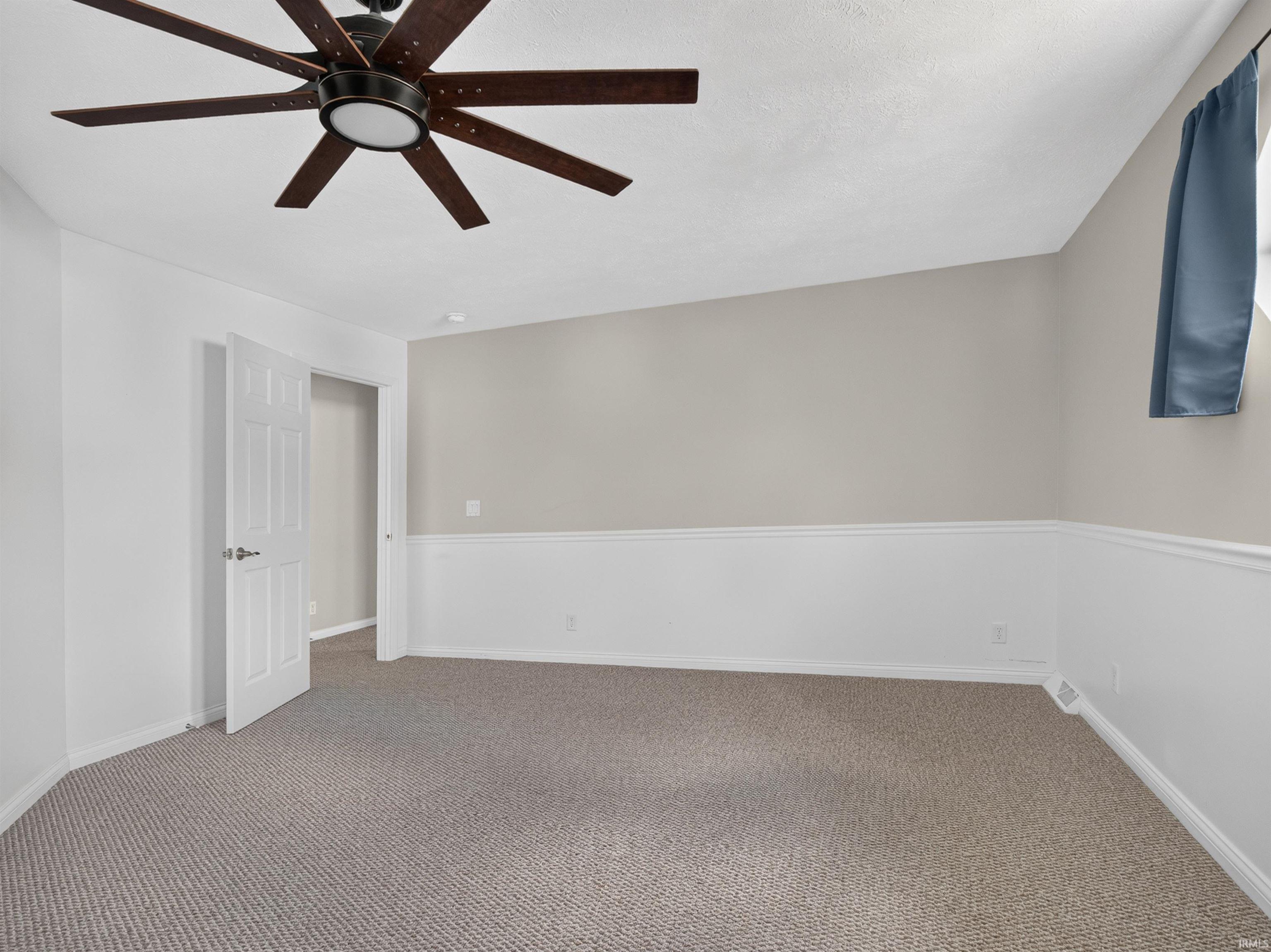 Spacious room featuring neutral carpeting, wainscoting, a multi-blade ceiling fan with integrated lighting, and a six-panel interior door