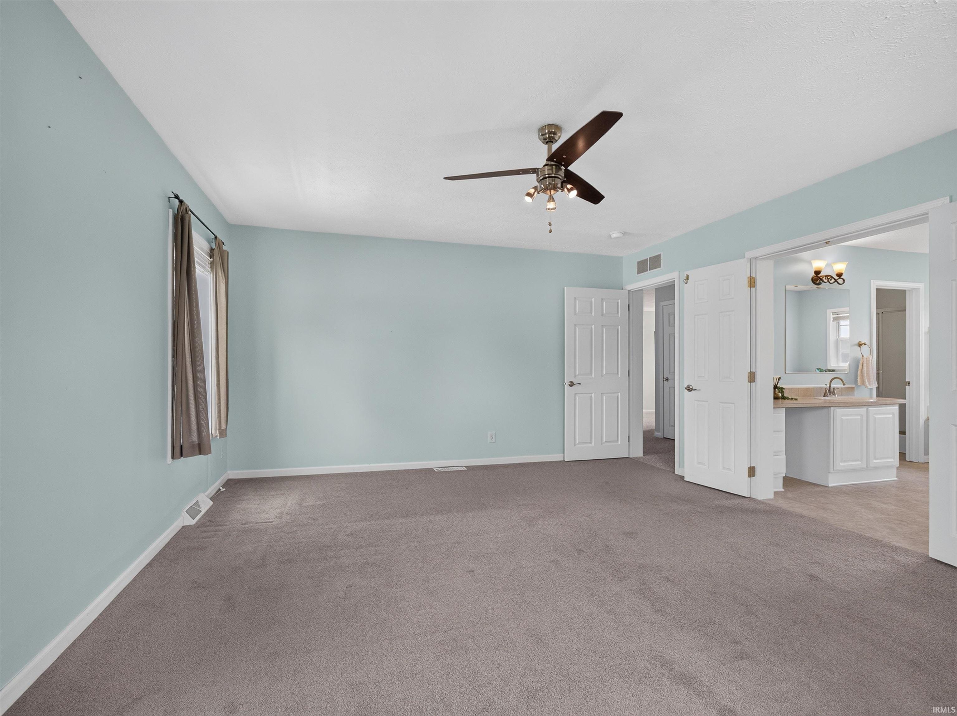 Spacious bedroom featuring light blue walls, grey carpet flooring, a ceiling fan with integrated lighting, and white baseboards