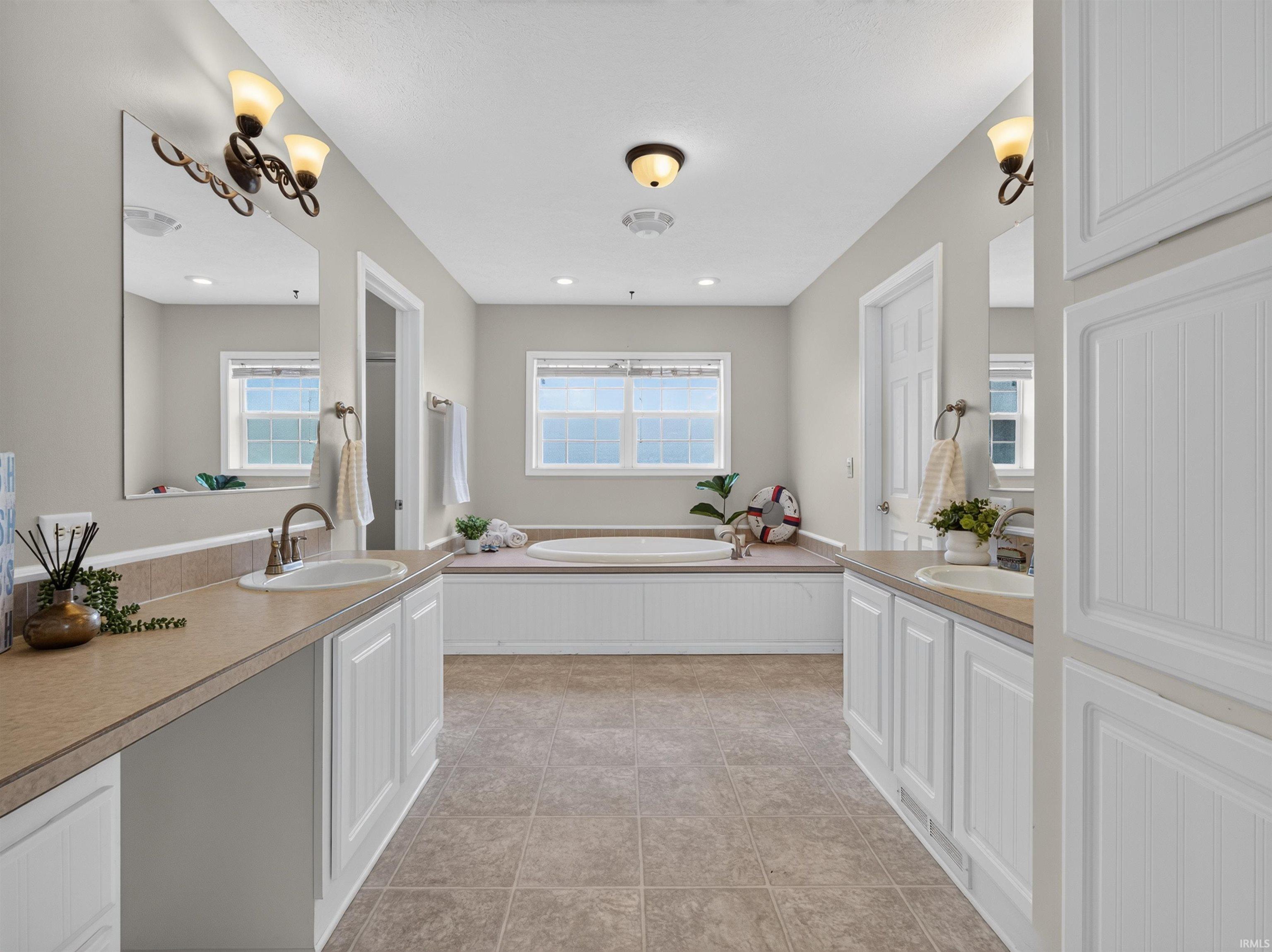 Spacious bathroom featuring a built-in soaking tub, dual vanity sinks, white cabinetry, two windows with natural light, and tile flooring