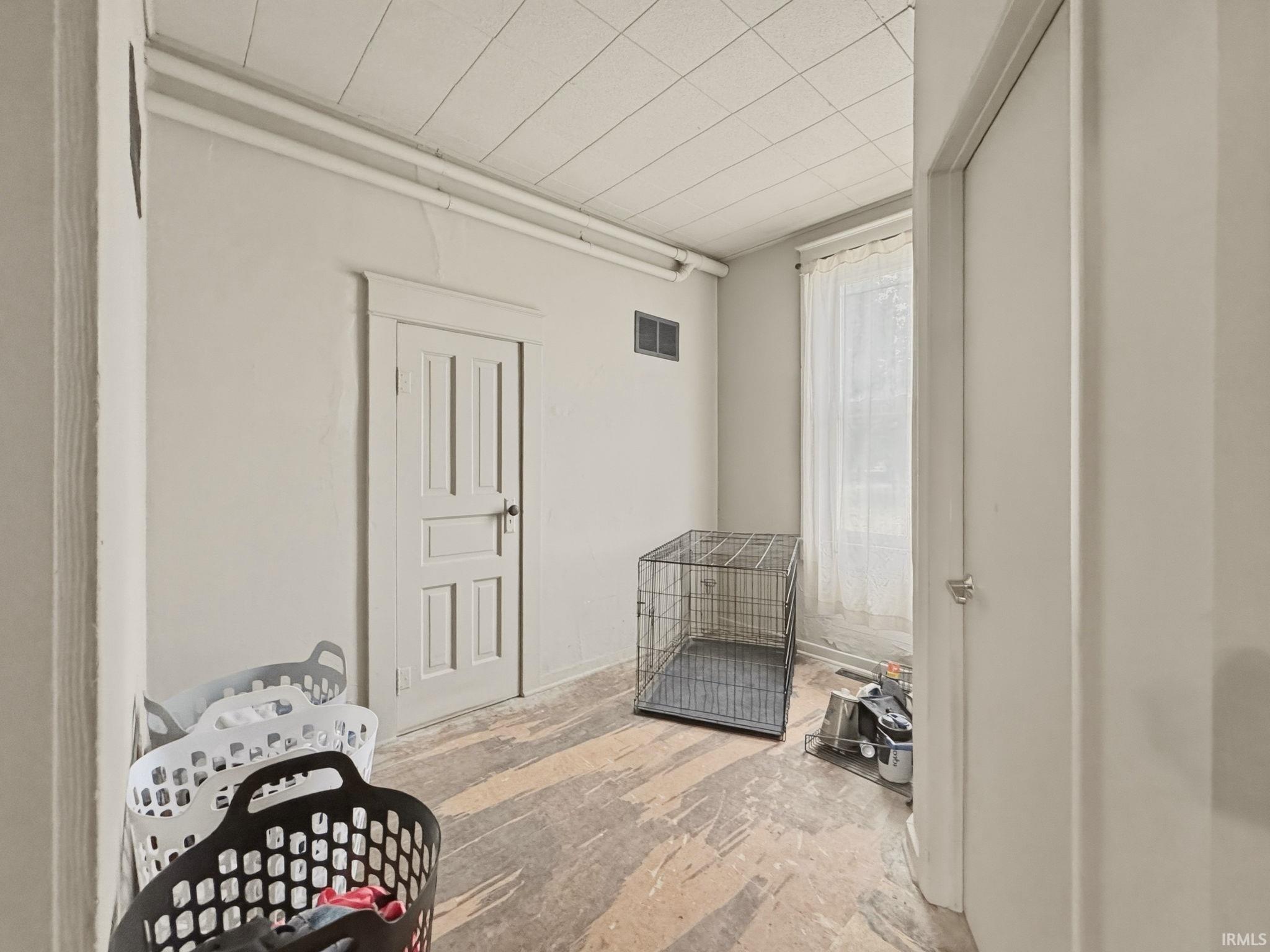 Interior space featuring a paneled door, a window with sheer curtains, a dropped ceiling, and exposed subflooring