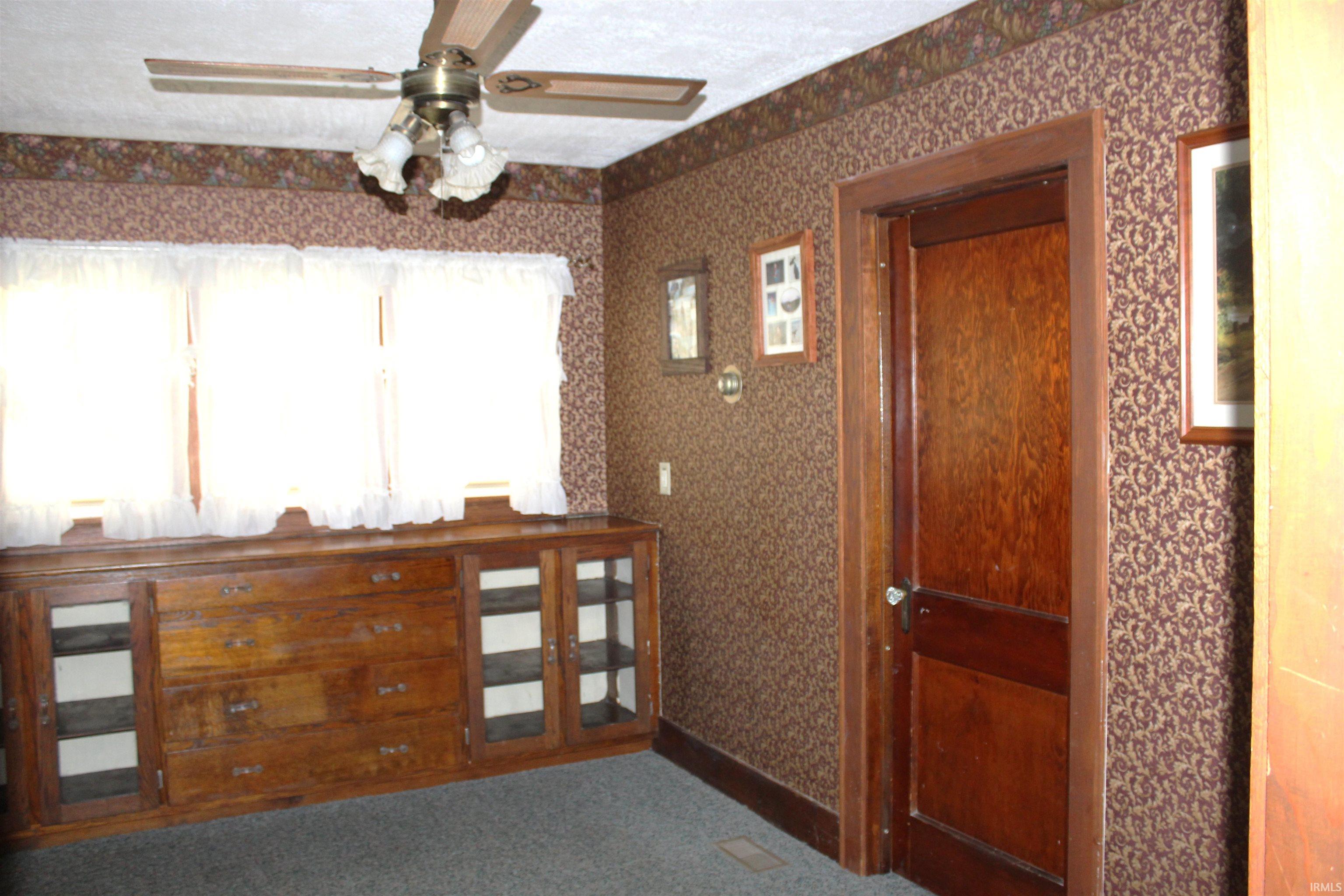 Built-in wooden cabinetry with glass-front storage, three windows featuring valance curtains, patterned wall coverings, a ceiling fan with integrated lighting, and a wooden interior door