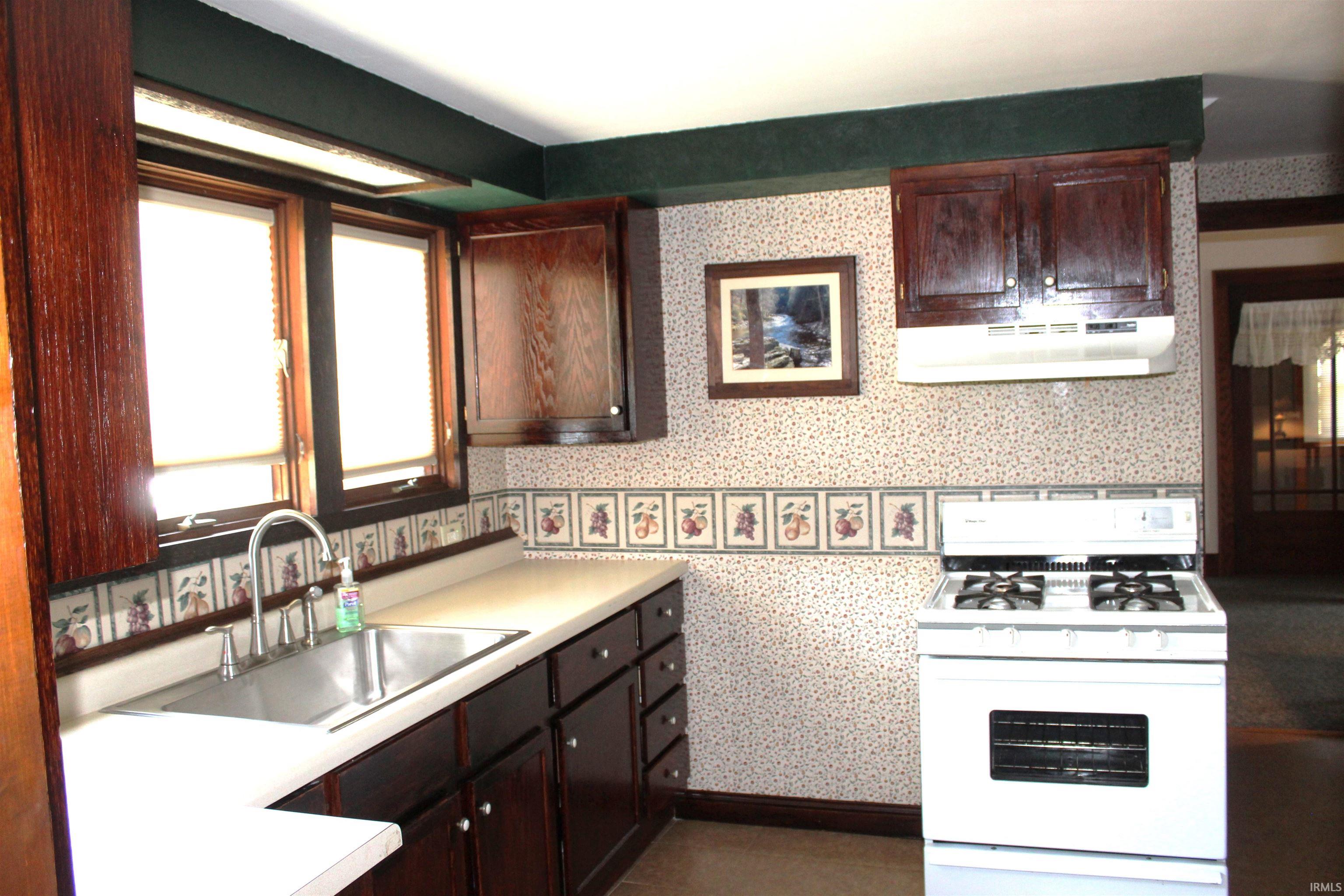 Kitchen featuring dark wood-finish cabinetry, a stainless steel sink with a chrome gooseneck faucet, light-colored countertops, a white freestanding range, and a window with roller shades