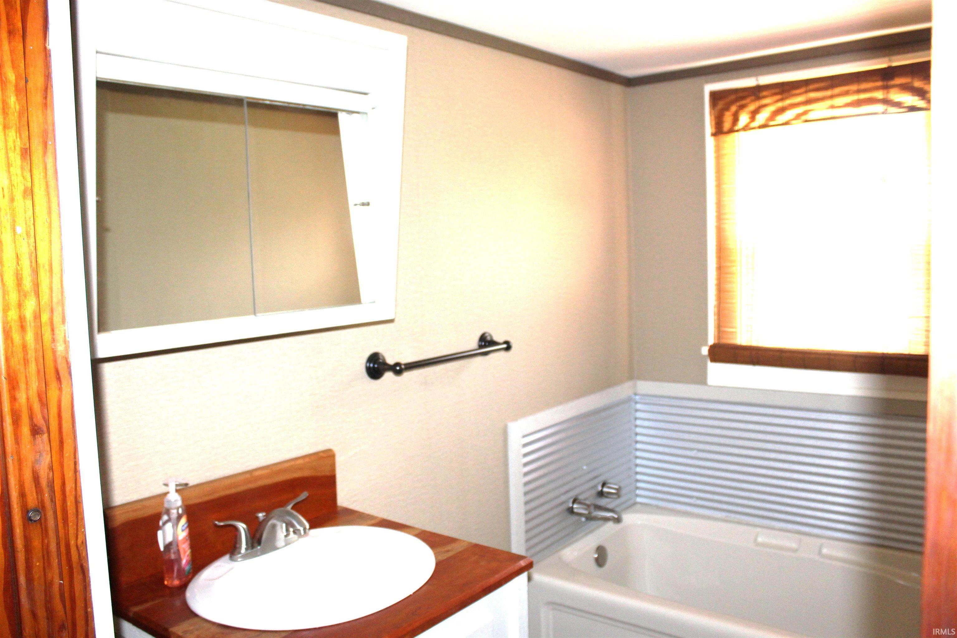 Bathroom featuring a white vanity cabinet with a wood-finish countertop, an oval undermount sink, a medicine cabinet, a bathtub with corrugated metal wainscoting, and a window with a woven shade