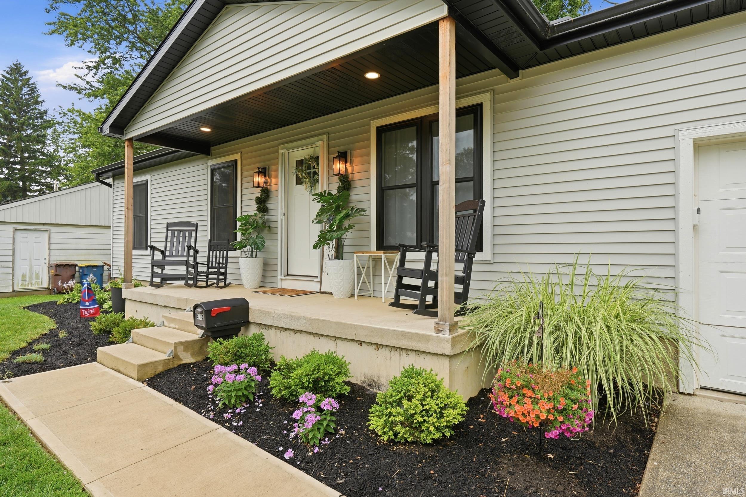 Front exterior featuring light gray siding, a covered porch with dark wood-finish support posts, and black exterior light fixtures flanking the entry