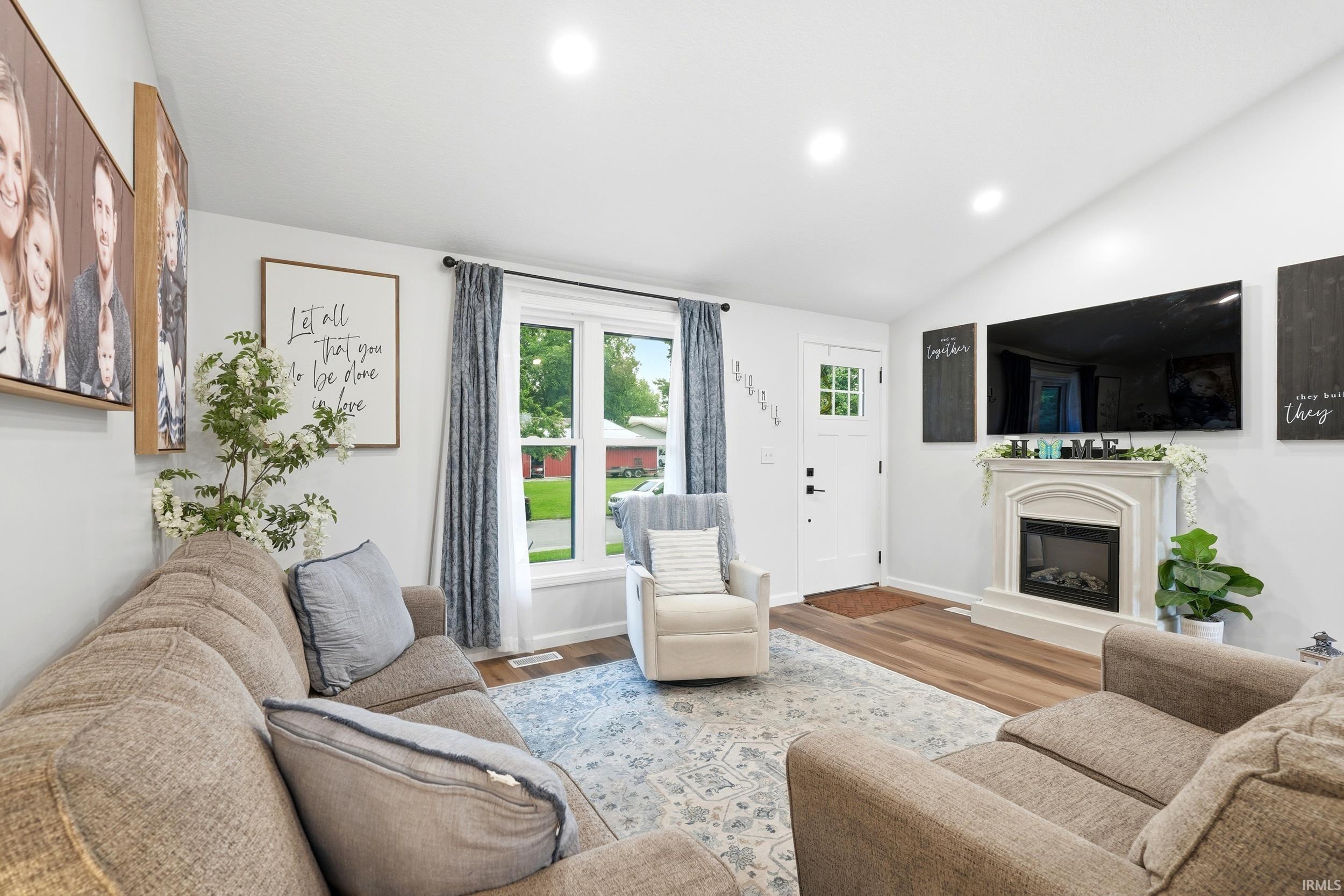 Living area featuring vaulted ceilings, recessed lighting, wood-finish flooring, a white paneled entry door with windowpanes, and a decorative fireplace with a mantel