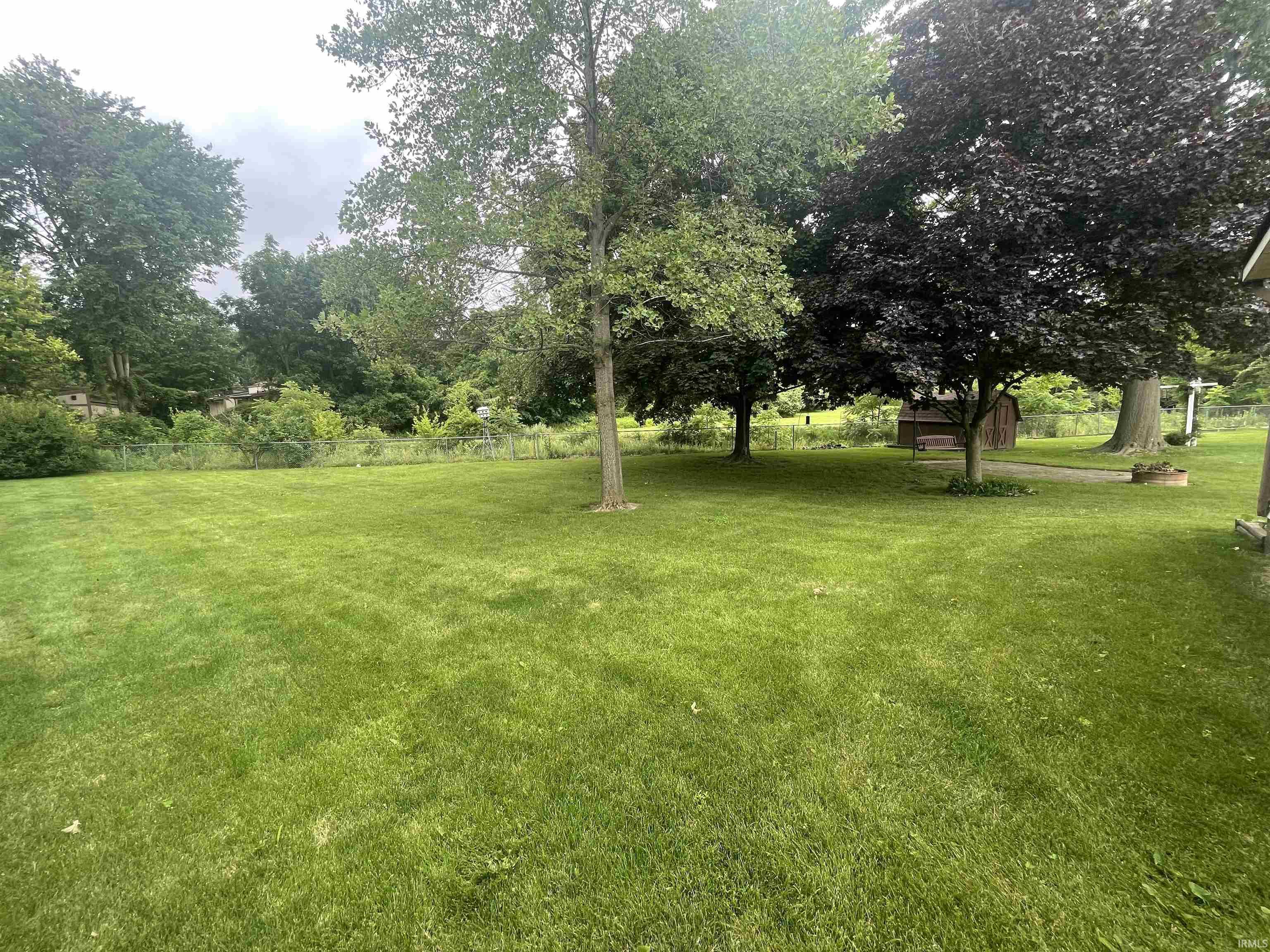 Expansive lawn area featuring mature trees, a chain-link fence, and a distant storage shed