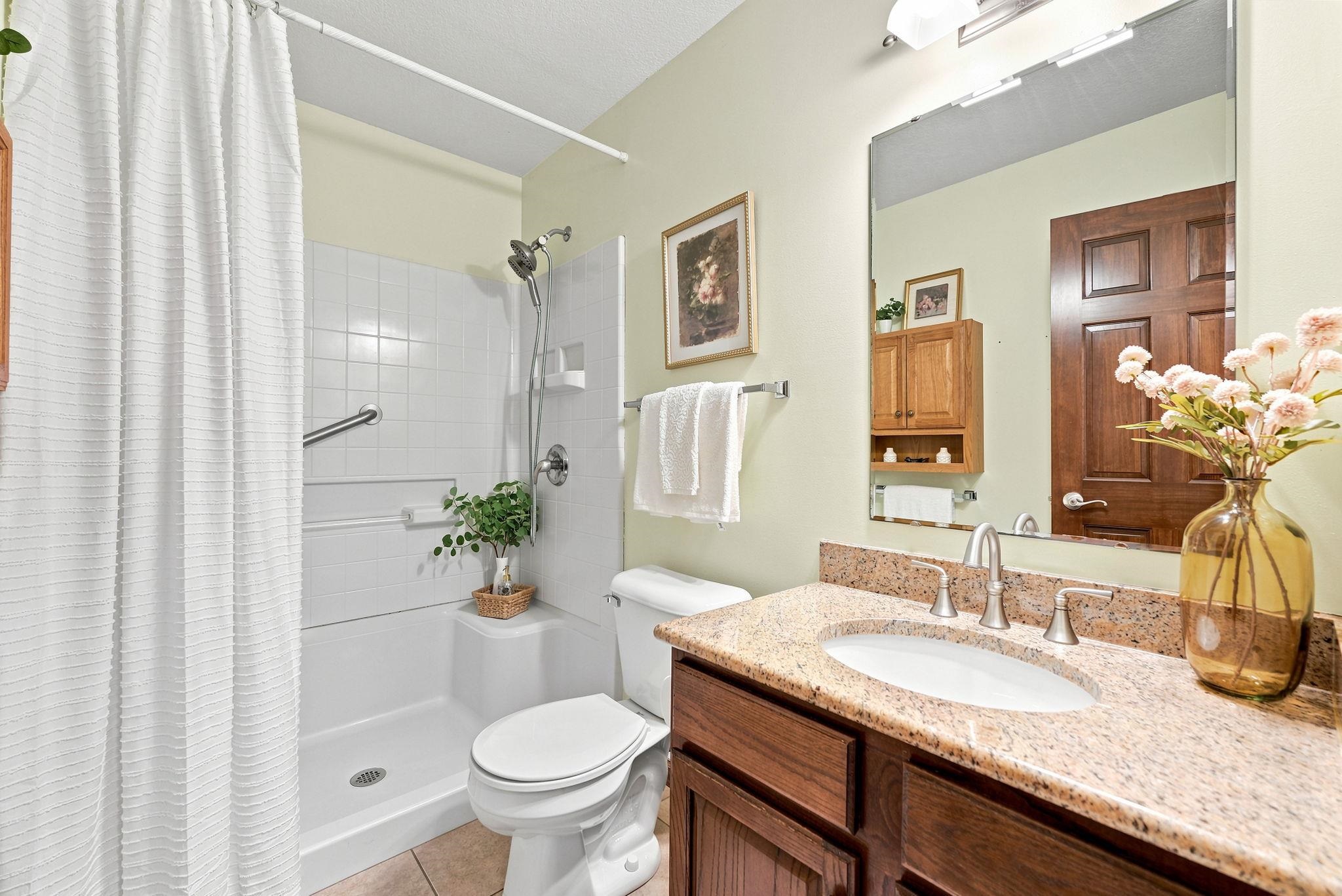 Bathroom featuring a walk-in shower with a grab bar, a single vanity with a granite countertop, a rectangular mirror, and wood-finish cabinetry