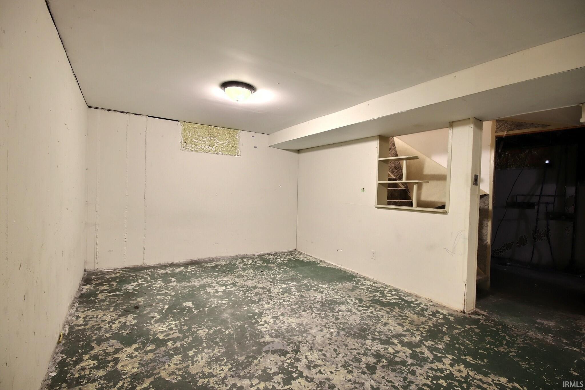 Finished basement space featuring a ceiling-mounted light fixture, painted concrete walls, and a built-in open shelving unit under the stairs