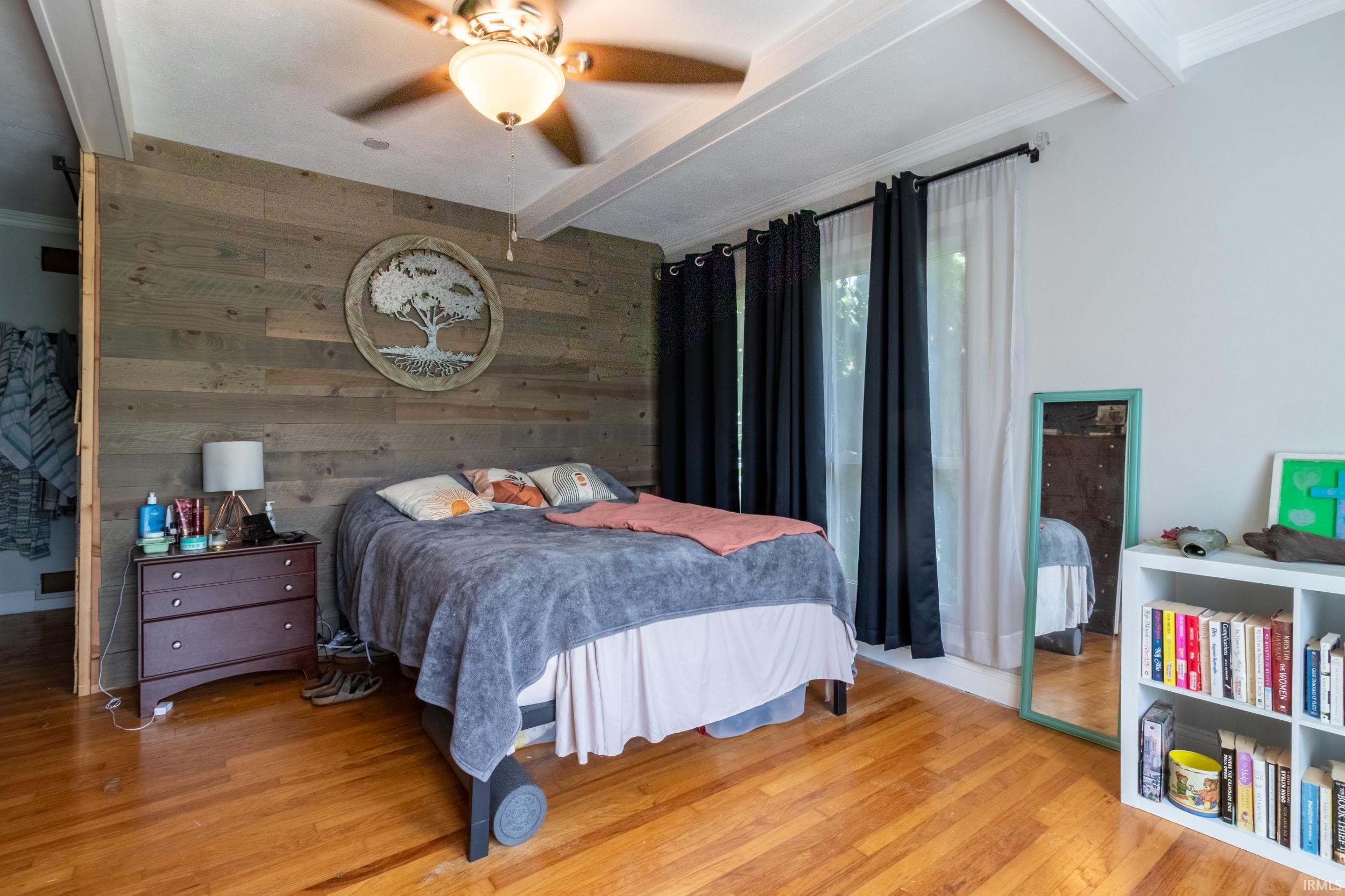Bedroom featuring a decorative wood-finish accent wall, wood-finish flooring, a ceiling fan with integrated lighting, and white ceiling beams