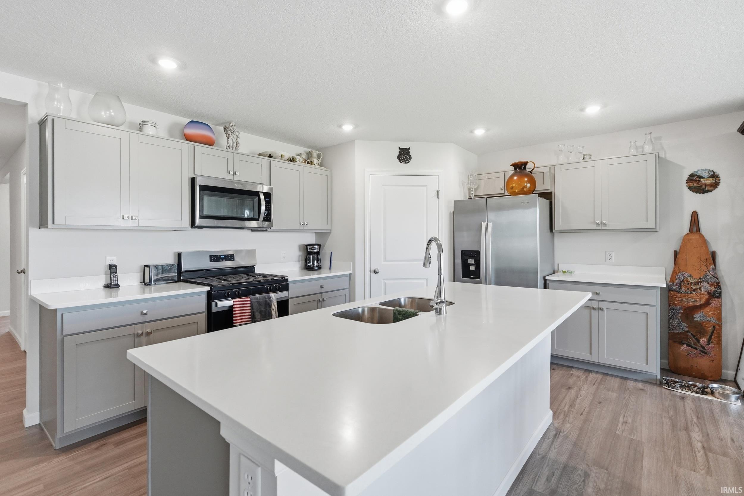 Kitchen featuring a large central island with double stainless steel sinks, white countertops, gray cabinetry, stainless steel appliances, and wood-finish flooring