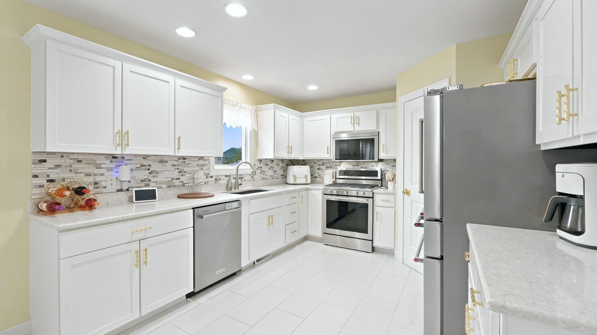 Kitchen featuring white shaker-style cabinetry with gold-tone hardware, stainless steel appliances, light-toned countertops, and a mosaic tile backsplash