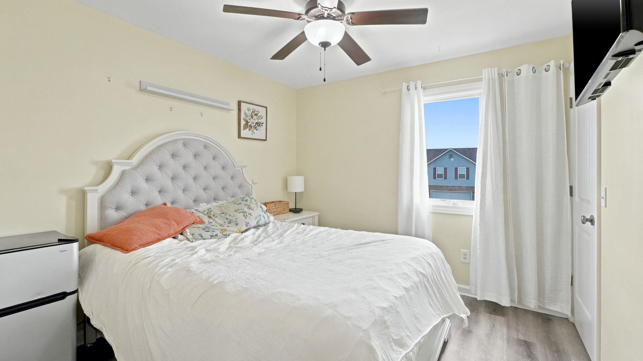 Neutral-toned room featuring wood-finish flooring, a ceiling fan with integrated lighting, and a window with white drapes