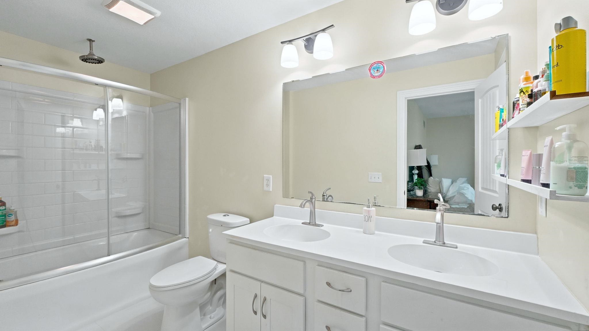 Bathroom featuring a double vanity with white cabinetry and integrated sinks, a full-width mirror, a shower-tub combination with sliding glass doors, and a ceiling-mounted rainfall showerhead