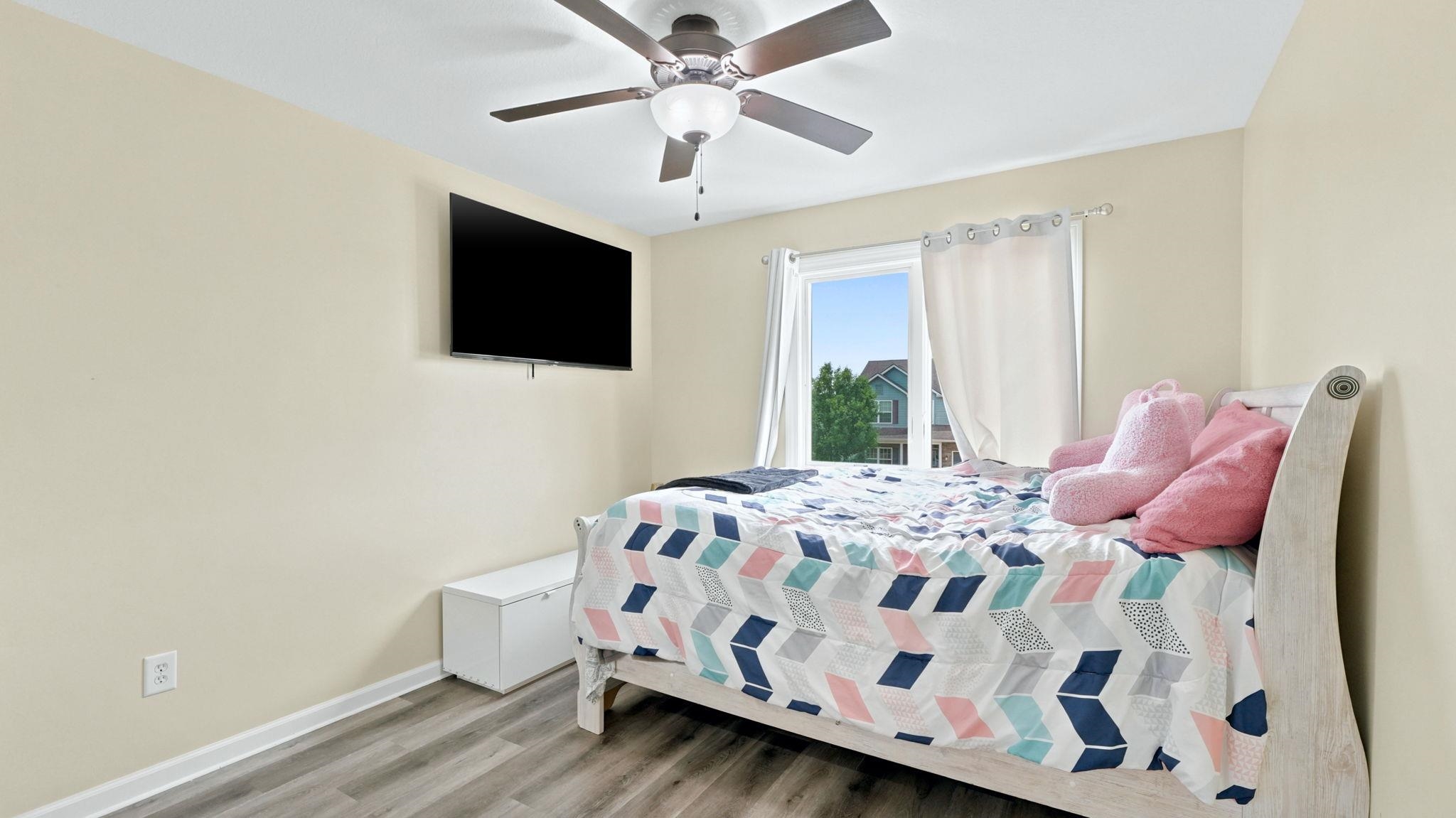 Bedroom featuring light-toned walls, wood-finish flooring, a ceiling fan with integrated lighting, and a window providing natural light