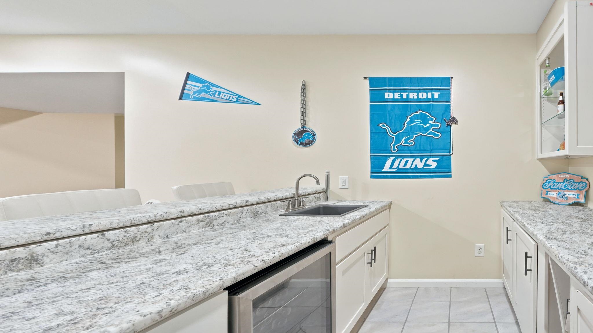 Built-in wet bar featuring a stainless steel sink, under-counter beverage refrigerator, white cabinetry with dark hardware, and granite-finish countertops