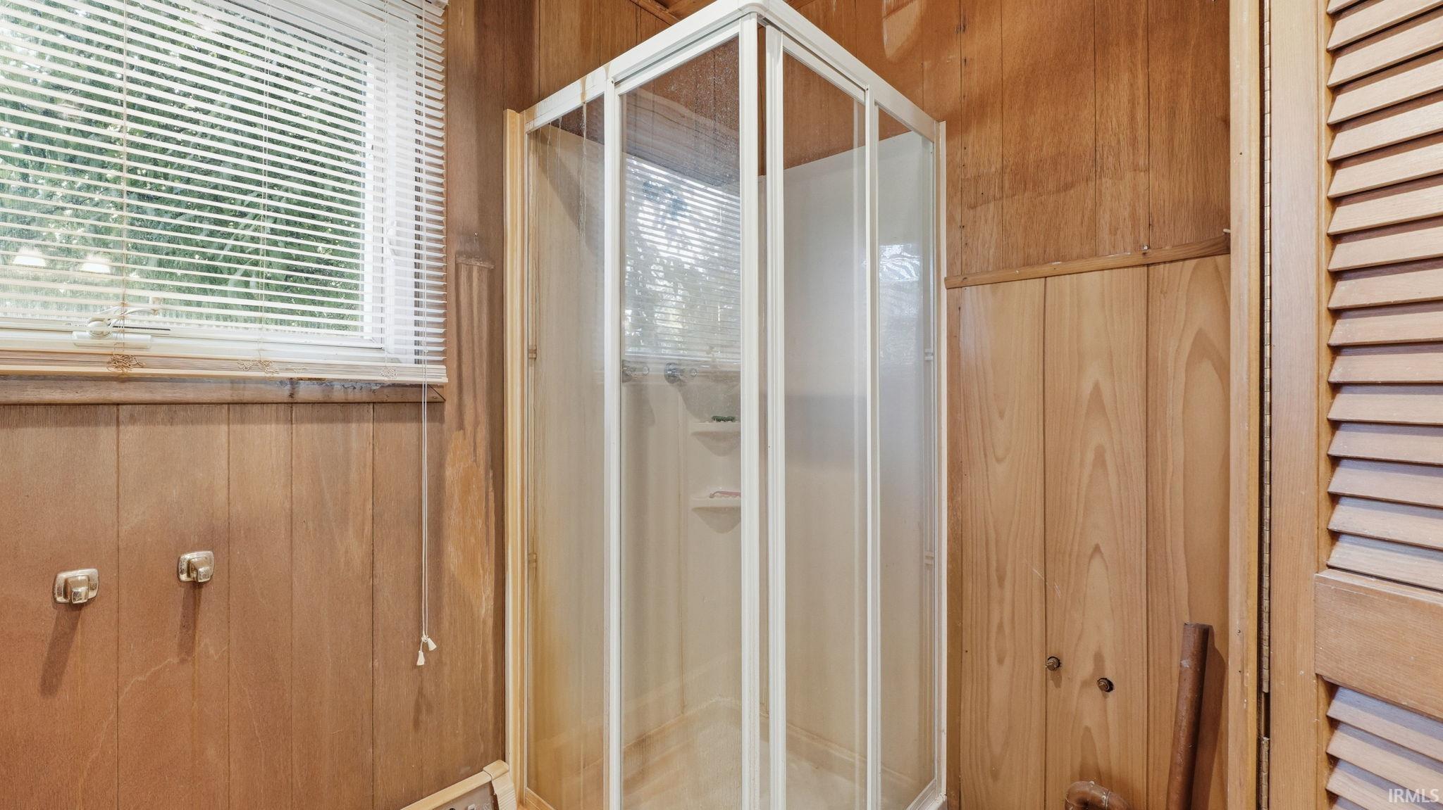 Corner shower enclosure with white framing, wood paneling, and a window with white blinds